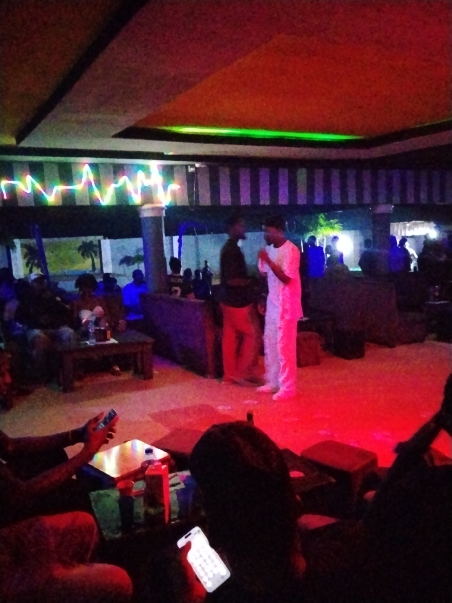 The image captures an indoor gathering at what appears to be a nightclub or lounge in Durumi, Nigeria. The scene is dimly lit, with vibrant neon lights and red lighting illuminating the space. In the foreground, two individuals are visible, one holding a smartphone with a brightly lit screen, and another person's hands are visible, also holding a phone. Further back, two men are engaged in a conversation; one is wearing a black shirt and orange pants, while the other is dressed in a white outfit. Other people are seated around tables, some with drinks on them. The environment features a patterned ceiling with colorful lights, striped walls, and dark furniture. The overall atmosphere suggests a social event or a night out.