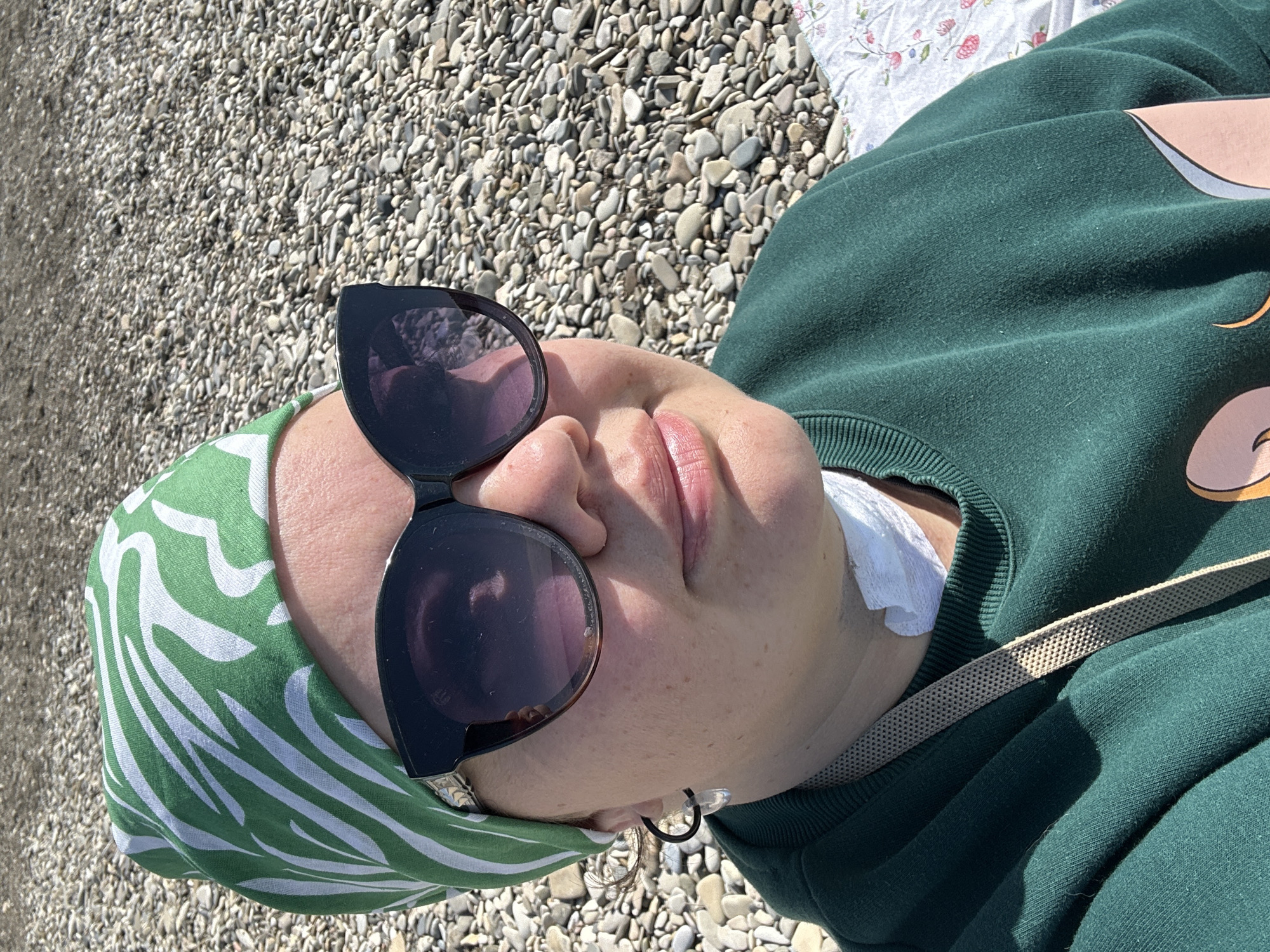 A person is lying down on a pebble-covered surface, possibly a beach, wearing a green and white patterned headscarf, sunglasses, and a green sweatshirt with a beige strap visible. A white bandage is visible on the person's neck. The location appears to be outdoors with bright sunlight. The image may have been taken in the city of Novomikhaylovsky, Russia, but it is difficult to verify this definitively without additional information.