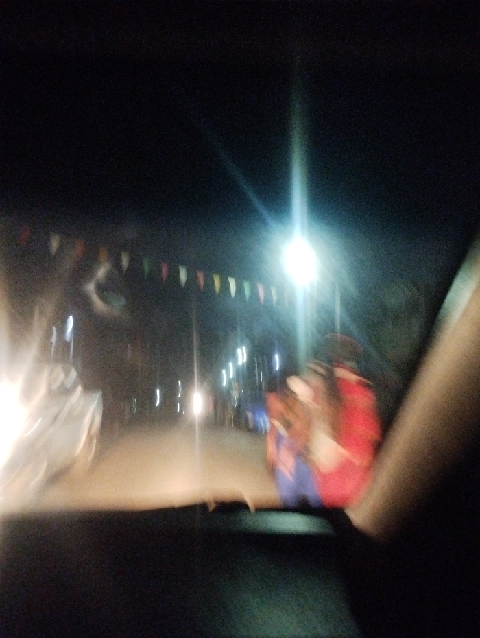 The photograph appears to be taken from inside a vehicle at night. There is a road ahead lit by streetlights and car headlights, giving off a bright glare. There are pennant flags strung along the road. The image is blurred, suggesting movement, and it's unclear if the colors are distorted. Passengers inside the vehicle appear to be wearing a red and blue top.