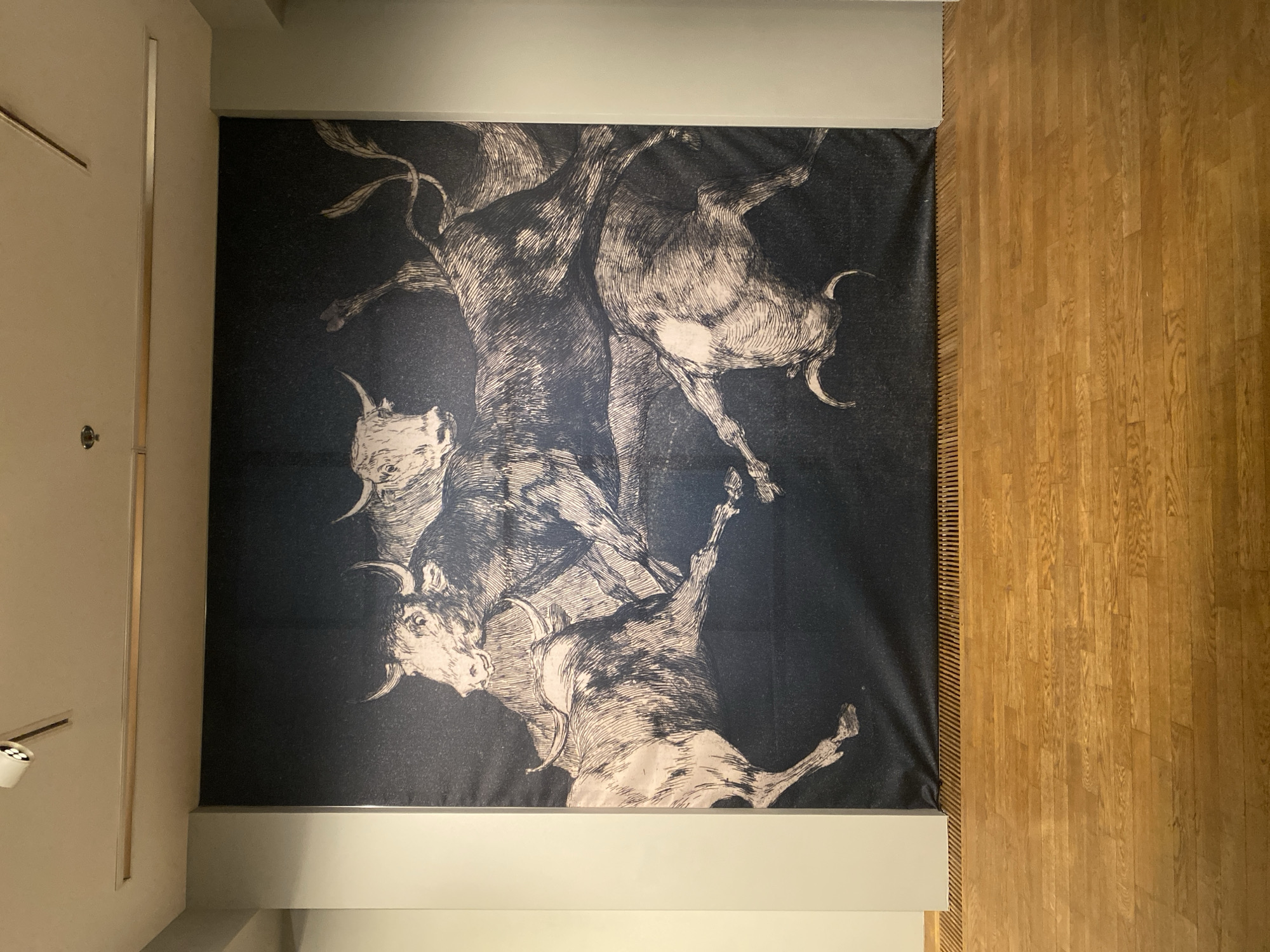 An expansive, monochrome tapestry depicting a dynamic scene of four bulls locked in a struggle dominates the wall of a modern gallery space. The bulls are rendered in a detailed, etched style against a dark, textured background. One bull in the foreground is on its hindquarters, its head low with horns pointed forward. To its left, another bull charges, its head raised and mouth open. Behind these, two more bulls are intertwined, their bodies and horns creating a chaotic and powerful composition. The tapestry is hung against a light gray wall, bordered by narrow white wall sections on either side. Above the tapestry, a track lighting system with multiple spotlights is installed on the off-white ceiling. Below the tapestry, a wooden floor with a polished, warm-toned finish is visible, interspersed with a dark metal grate running along the base of the wall. The image was taken at the National Gallery - Palace, located at 1 "Knyaz Alexander I" Square in Sofia, Bulgaria.