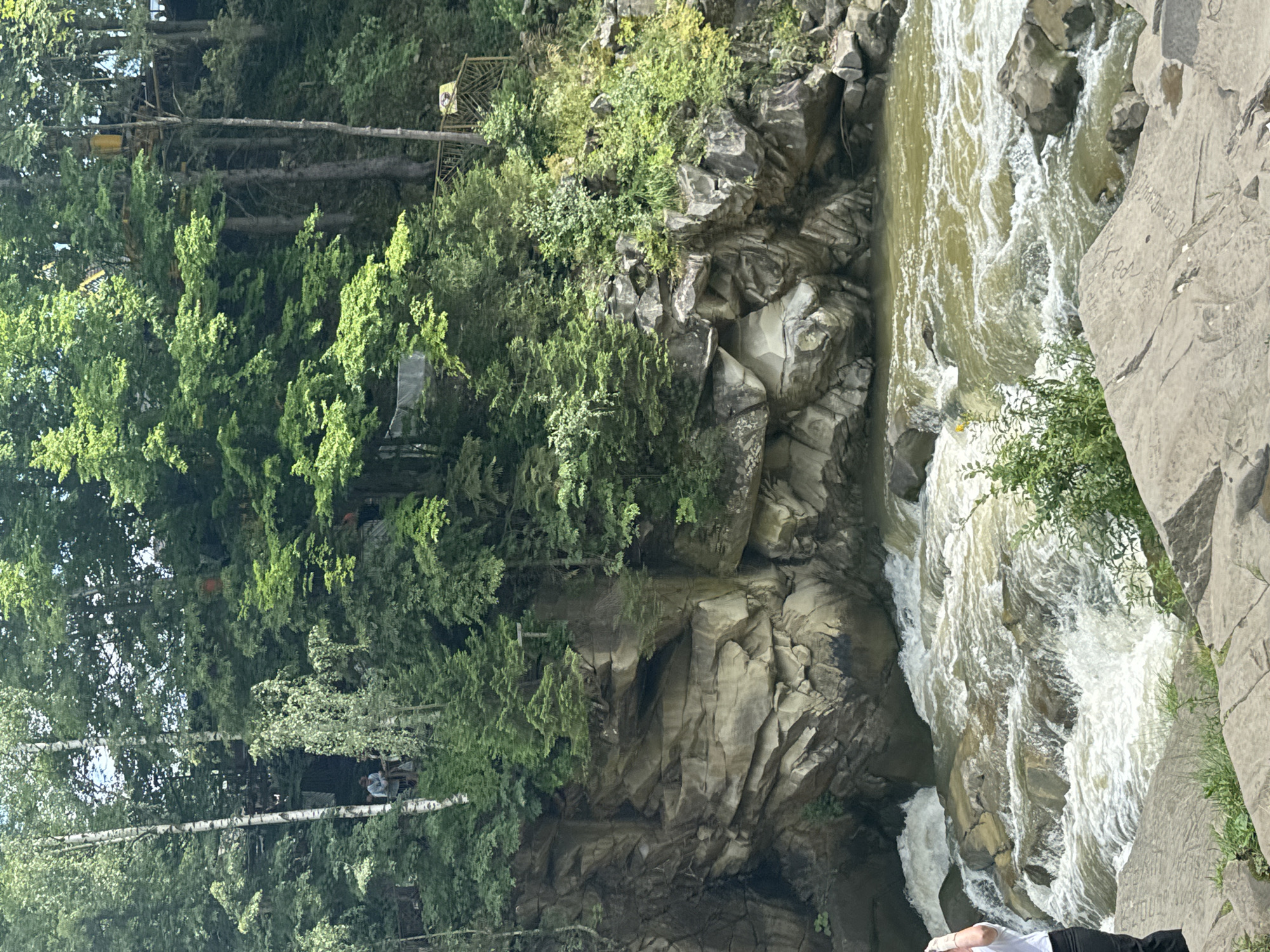 The image depicts the Prut River flowing through a rocky gorge in Yaremche, Ukraine. The water is turbulent, with white rapids cascading over numerous stones. On the left side of the river, a steep, rocky cliff face rises, covered in patches of green vegetation. On the right bank, the gorge continues with more rocks and dense green trees. High above the gorge, nestled within the trees, are some man-made structures. These include what appear to be platforms or walkways built among the trees, with hints of yellow and wooden elements. One cropped image shows a yellow structure that resembles an observation deck or a part of an adventure park. In the distance, on a higher level, a person can be seen standing on a wooden platform, surrounded by trees. Another cropped image shows a glimpse of a wooden staircase leading up the embankment on the right side. The overall environment suggests a natural landscape with recreational facilities integrated into the forest. The time of day appears to be daytime, with diffused sunlight filtering through the tree canopy.