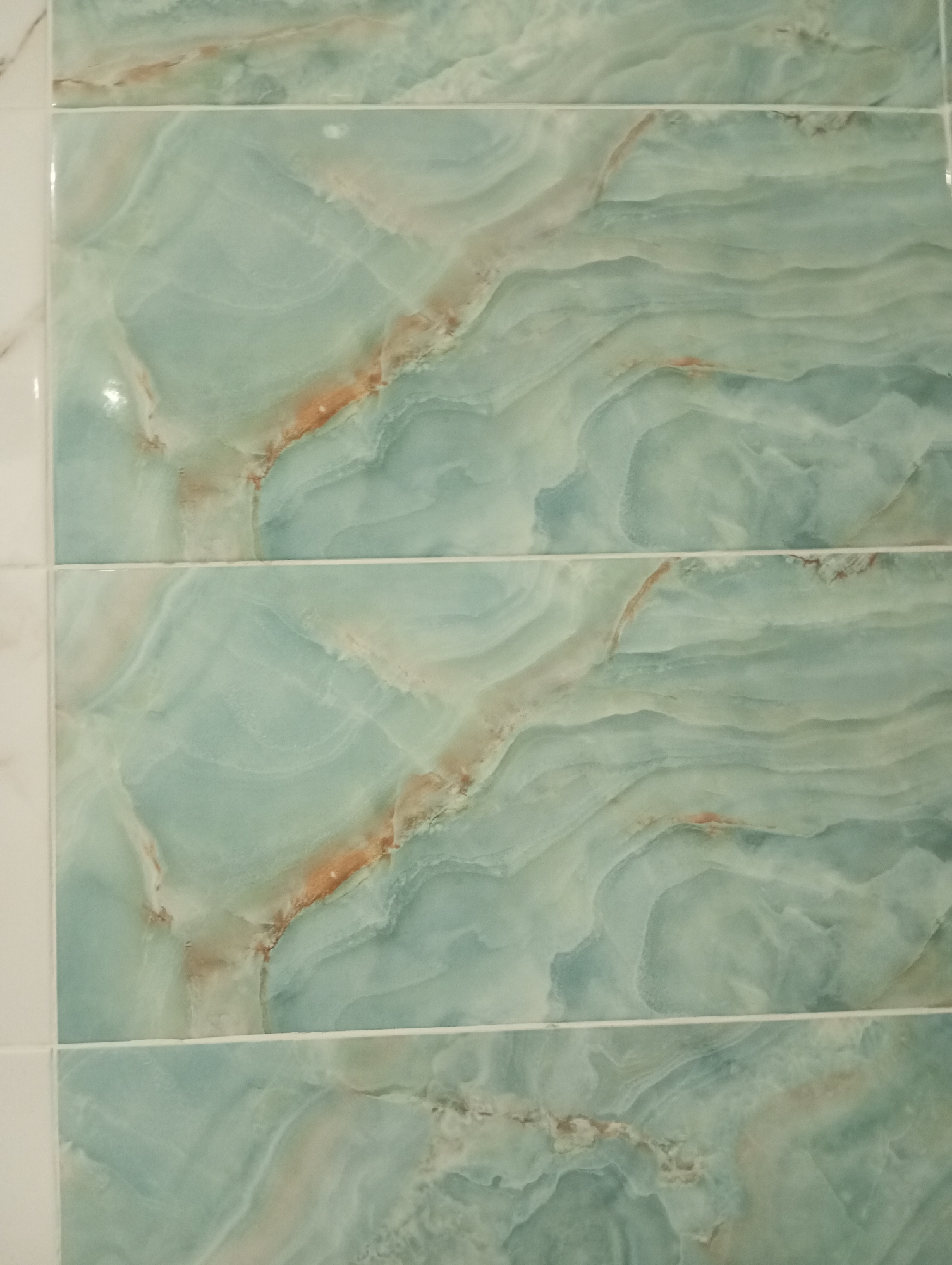 The image displays a close-up view of a tiled wall, likely in a bathroom or kitchen. The tiles are rectangular and have a marbled pattern with swirling shades of teal, white, and a reddish-brown hue. The grout lines between the tiles are thin and white. The lighting suggests an indoor setting, with a slight sheen reflecting off the tile surfaces. There are no people, objects, or actions visible in the frame. The image does not contain any information that would allow for the identification of its specific location within Verkhnyaya Pyshma, Russia.