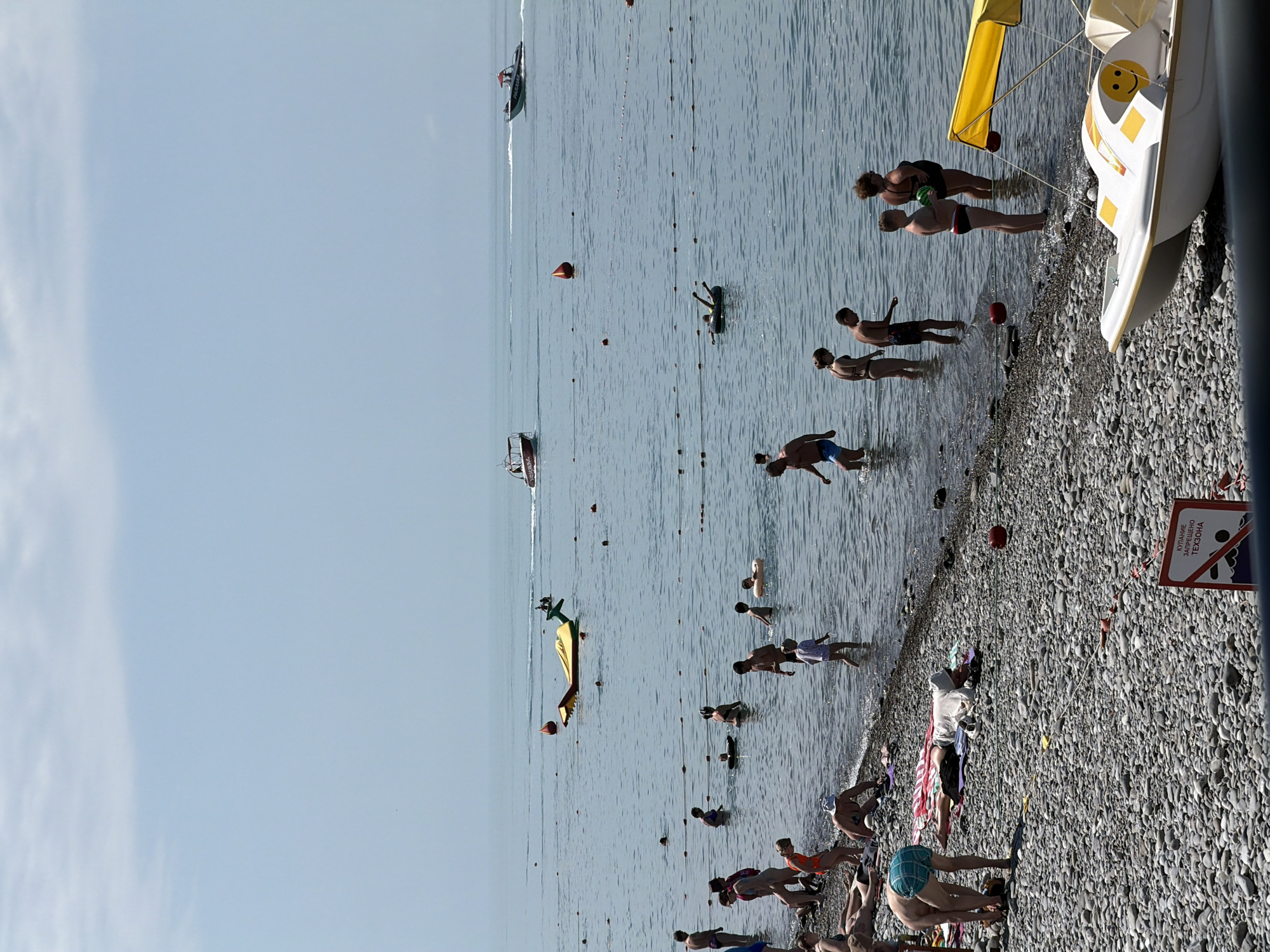 This is an image taken on a sunny day at a crowded beach in Sochi, Russia, likely along the Black Sea coast. The foreground shows a pebbled shoreline with several people sunbathing and relaxing on towels. A yellow and white pedal boat with a smiley face design is docked on the beach.
The middle ground depicts the shallow waters of the sea, teeming with people wading, swimming, and playing. There are several inflatable yellow buoys and a rope barrier marking off sections of the water. In the distance, a yellow inflatable toy resembling a dragon is floating.
The background features the calm, blue expanse of the sea under a clear sky with scattered clouds. Several boats are visible on the water, including a speedboat and a smaller passenger boat. A red buoy is also present in the sea. A sign with Russian text, likely indicating a restricted area or a warning, is visible on the lower right side of the image. The overall scene conveys a typical summer vacation atmosphere at a popular seaside resort.