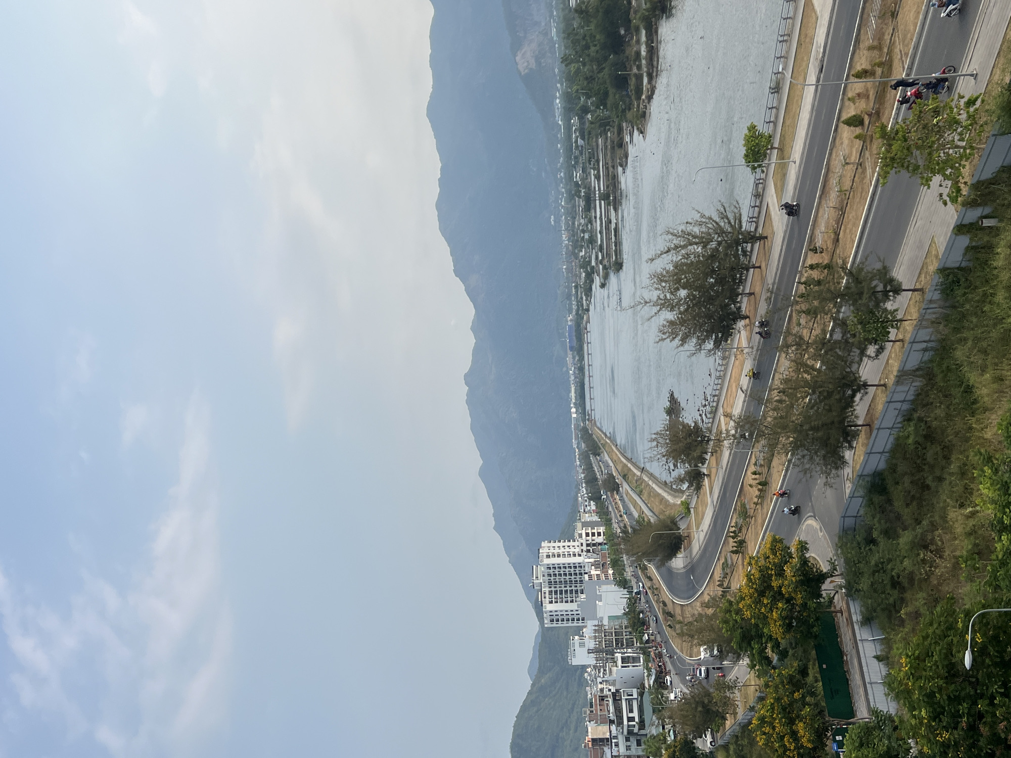 This high-angle image provides a panoramic view of a coastal or riverine landscape, likely in the Nam Natural Heritage Area of Trang, Vietnam. The scene is dominated by a majestic mountain range, a wide body of water, a winding road, and a cluster of buildings, all under a hazy sky.
In the mid-ground, a multi-lane road, possibly a coastal highway, curves and stretches parallel to the water. It features a central divider and is lined with trees and streetlights. Several motorbikes are visible on the road, indicating active daily traffic, with people riding them. To the right of the road, particularly in the foreground, dense green foliage, including trees with some yellow blossoms, fills the space.
To the left of the road lies a broad body of water, appearing calm and somewhat murky, possibly a river or a bay. Along its far bank or within the water, some structures or small vessels can be seen. Further across the water, a collection of modern, white multi-story buildings forms an urban or residential area nestled at the base of the mountains.
The background is dominated by a vast range of mountains, their slopes covered in lush, dark green vegetation. These mountains extend into the distant, hazy horizon, providing a dramatic backdrop to the scene. The sky above is a pale, hazy blue with subtle, indistinct white cloud formations, suggesting either an overcast day or atmospheric haze.
The image captures a daytime scene, characterized by diffused natural light. The overall impression is one of a busy yet scenic coastal environment, with the natural beauty of the mountains and water contrasting with the human development along the shore. No visible text is discernible in the image.