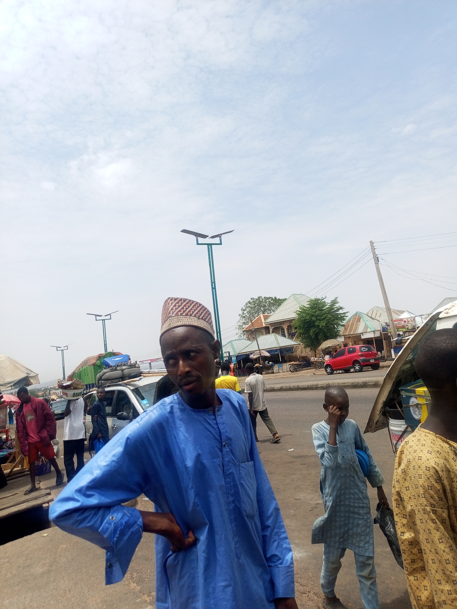 The image captures a street scene in Gombe, Nigeria. Several people are visible, including a man in the foreground wearing a blue traditional garment and a patterned cap. To his right, a child dressed in light blue clothing is standing with a hand on their face. Another person wearing a yellow patterned garment is partially visible on the right edge.
In the background, there are other individuals walking along the side of a road. Several vehicles are parked or moving on the road. Streetlights are positioned along the street, and buildings with corrugated metal roofs are visible in the distance. The sky is overcast. There are also some objects placed on top of a parked car and people carrying items on their heads.