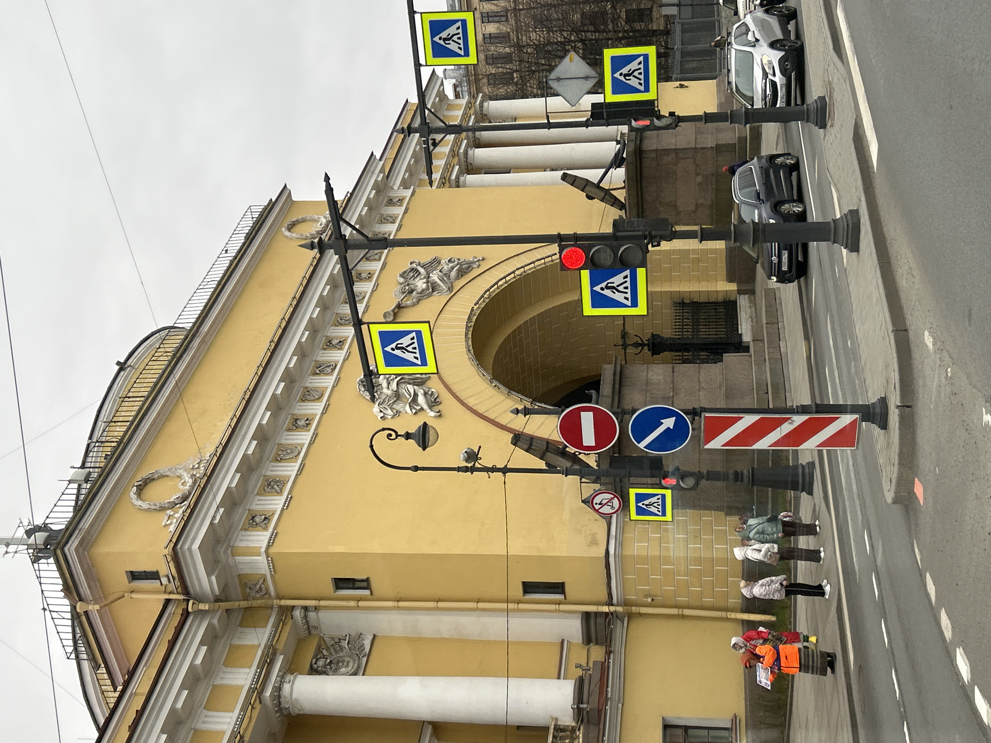 This image captures a vibrant street scene in Saint Petersburg, Russia, on an overcast day. The focal point is a grand, ornate yellow building with classical architectural elements, including large white columns, an arched entrance, and decorative relief sculptures featuring figures and garlands.
Along the street in front of the building, numerous traffic signs and lights are mounted on poles. Prominently displayed are multiple blue square signs with a white walking figure, indicating pedestrian crossings. Other visible signs include a red circular "no entry" sign, a blue circular "turn right" arrow sign, and a red and white striped warning sign. A traffic light displays a red signal. Utility wires span across the top of the frame against a muted, cloudy sky.
On the sidewalk, several pedestrians are present. Four individuals, dressed in winter coats, are standing together, possibly waiting to cross the street. Further down, another person in an orange high-visibility vest is visible. Various cars are on the street; a silver SUV with license plate "X 889 CY 178" and a black SUV with "A 233 ME 178" are clearly visible, with the "178" region code confirming the Saint Petersburg location. The overall atmosphere suggests a typical daytime city environment, with no discernible emotions from the subjects.