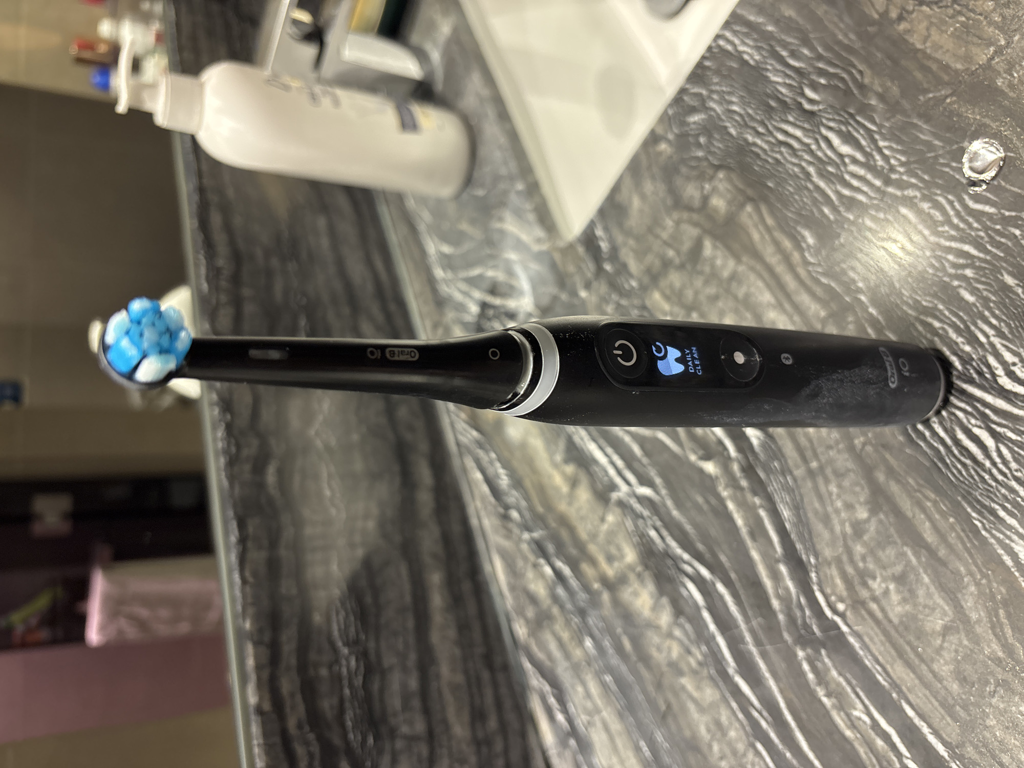 A black Oral-B electric toothbrush with a blue and white brush head stands upright on a textured, grey countertop. The toothbrush displays a digital screen showing a power button icon and a stylized tooth icon with the words "DAILY CLEAN" below it. To the right of the toothbrush, a white pump bottle of lotion or soap is visible. The background shows a blurred reflection in a mirror, with a glimpse of a bathroom sink. The scene is consistent with a personal bathroom counter in Dubai, United Arab Emirates.