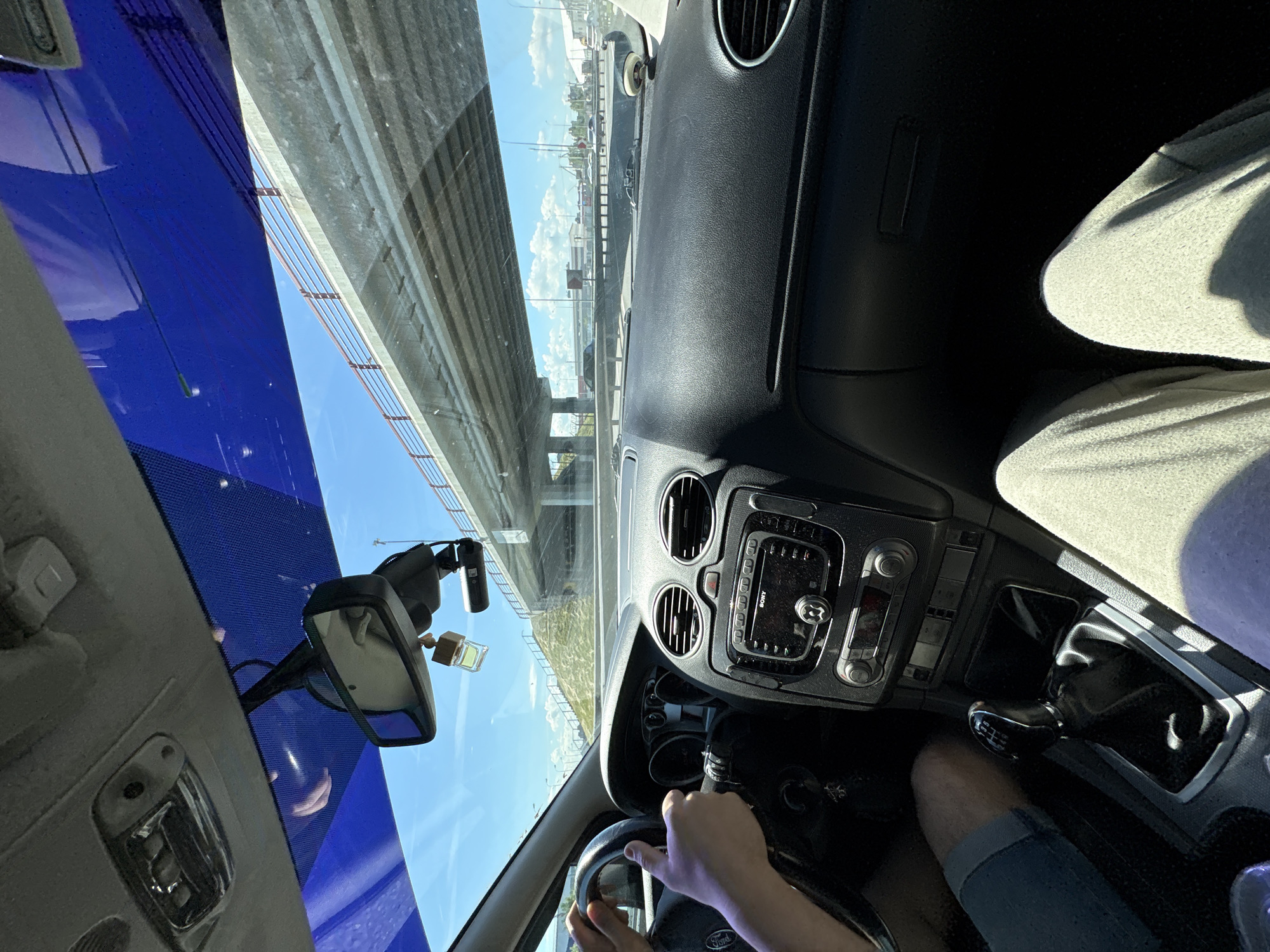 The image shows the interior of a car, viewed from the passenger seat, with a driver's hands on the steering wheel. The car is on a highway, passing under an elevated road. The scene appears to be in Novosaratovka, Russia, given the context provided. The driver is wearing a blue shorts and the passenger is wearing light gray sweatpants. The dashboard features a Sony audio system, air vents, and a gear shift. A rearview mirror with a dashboard camera and an air freshener hangs from the windshield. The sky is blue with some clouds, and sunlight is casting shadows. The elevated road has guardrails and supports, and the surrounding landscape includes grassy slopes and other vehicles on the highway.