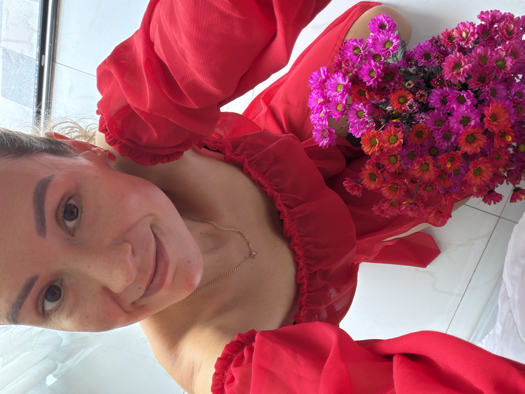 A young woman in a red off-the-shoulder dress kneels on a white tiled floor, holding a bouquet of pink and orange chrysanthemums. She looks up at the camera with a slight smile, her blonde hair pulled back. The background shows a bright, indoor environment with a hint of outdoor light filtering in. The woman appears to be in a residential setting, possibly an apartment or house, in Nha Trang Ward, Vietnam, suggested by the typical tiling and natural lighting conditions found in that region.