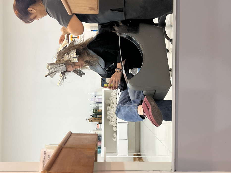 The image captures an indoor scene within a hair salon in Puebla, Mexico. Two individuals are present. A young adult female client, with numerous sections of her hair wrapped in aluminum foil, sits in a gray salon chair. She wears a black salon cape over a dark top and blue jeans, with maroon platform shoes. Her attention is directed downwards towards a yellow mobile phone held in her right hand. A plastic water bottle rests on the armrest of her chair. Behind her, an adult female hair stylist, dressed in a dark uniform, is actively working on the client's hair, adjusting or applying product to the foiled sections. The environment features white walls and a white tiled floor. To the left, a white shelving unit holds various hair product bottles and neatly rolled white towels. A wooden cabinet is partially visible on the left, with a paper displaying text including "INFINITUM6RDZ" and "Ca9i." The lower portion of the scene is a reflection in a large mirror.