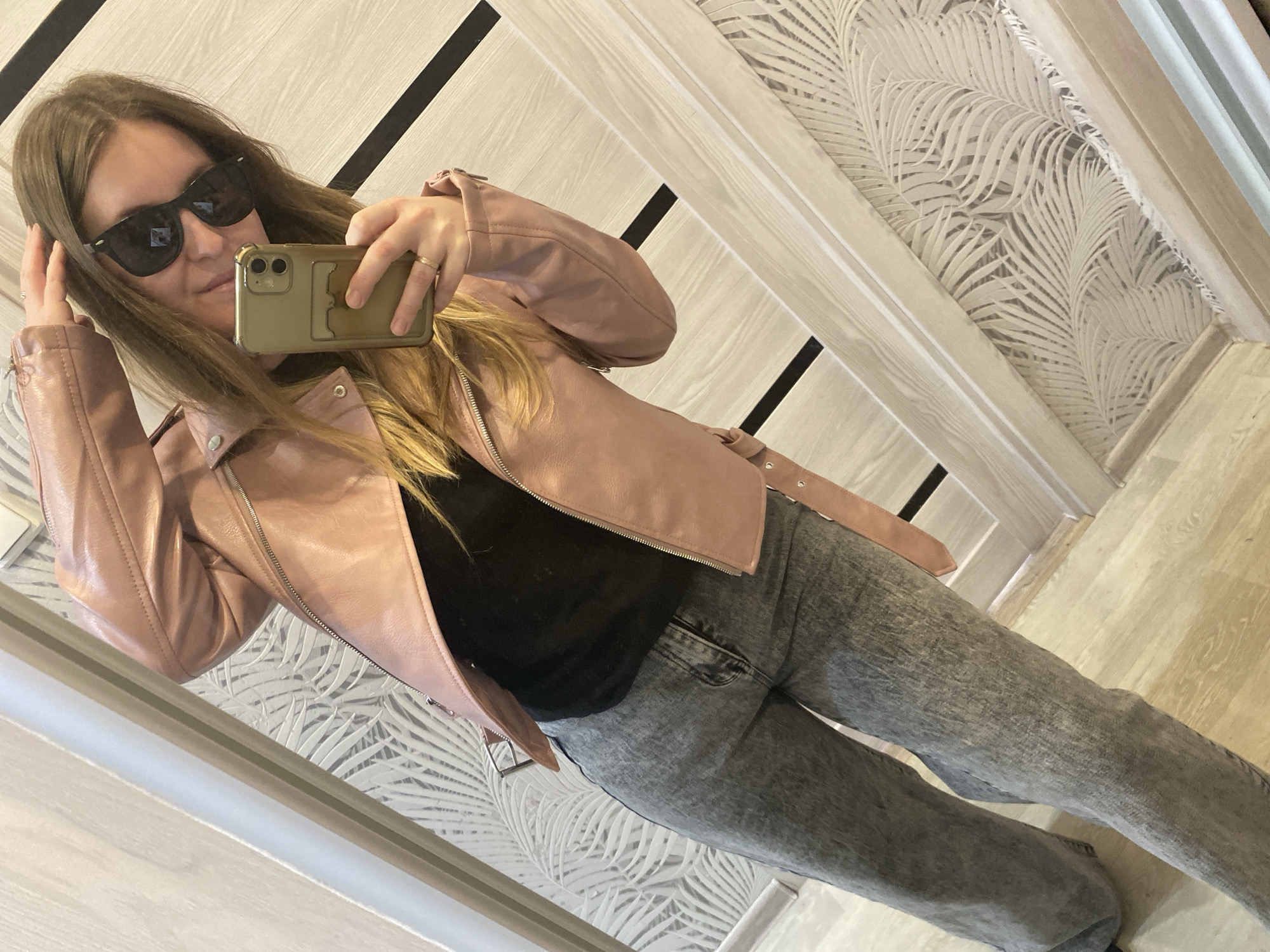 A young, light-skinned woman with shoulder-length blonde hair and wearing sunglasses is taking a selfie in what appears to be a room. She is wearing a pink leather jacket over a black top and gray jeans. She is holding her phone up to take the picture. The background includes a patterned wallpaper featuring palm leaves and wood paneling on the door. The location cannot be determined.