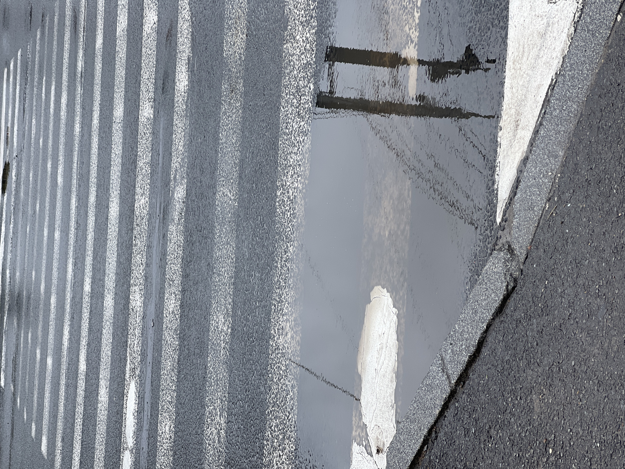 The image shows a wet pedestrian crossing. The crossing stripes are white and parallel, painted on dark asphalt. A large puddle of water covers a portion of the crossing, reflecting a sky with clouds and two vertical structures, likely poles or posts. The curb is made of grey, textured stone. The overall scene suggests a rainy day in an urban environment, though no specific city or landmarks are identifiable.