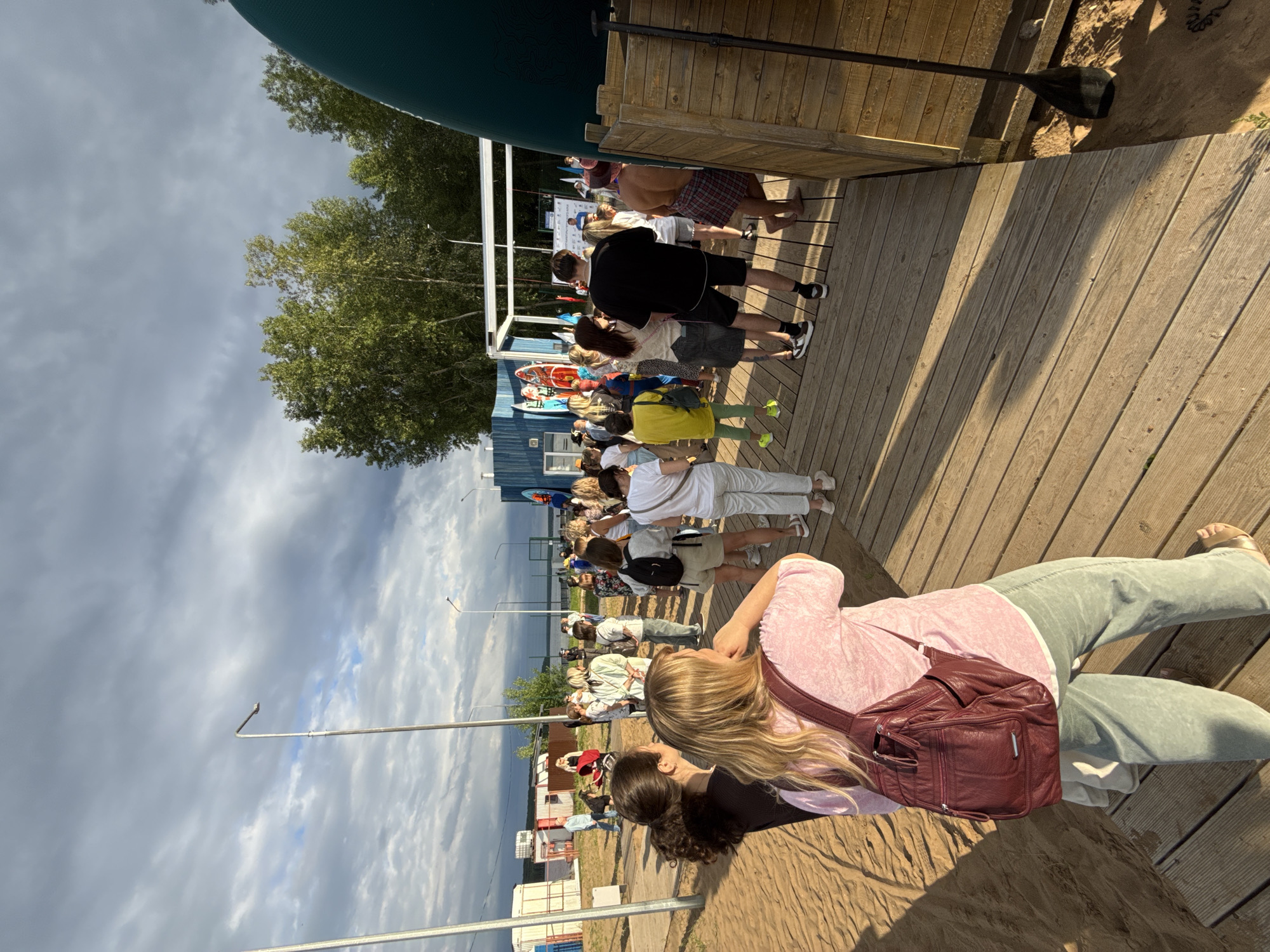 The image depicts an outdoor gathering on a sandy area with a wooden boardwalk. A crowd of people, dressed in casual summer attire, are standing and milling about. In the background, there are some modular buildings, trees, and a body of water. A stage or platform area is visible with people standing on it. Several surfboards or paddleboards are leaning against a blue structure. The sky is overcast with dramatic clouds. The scene appears to be a public event or festival, possibly at a waterfront location. The provided information specifies the location as Chaikovsky, Russia.