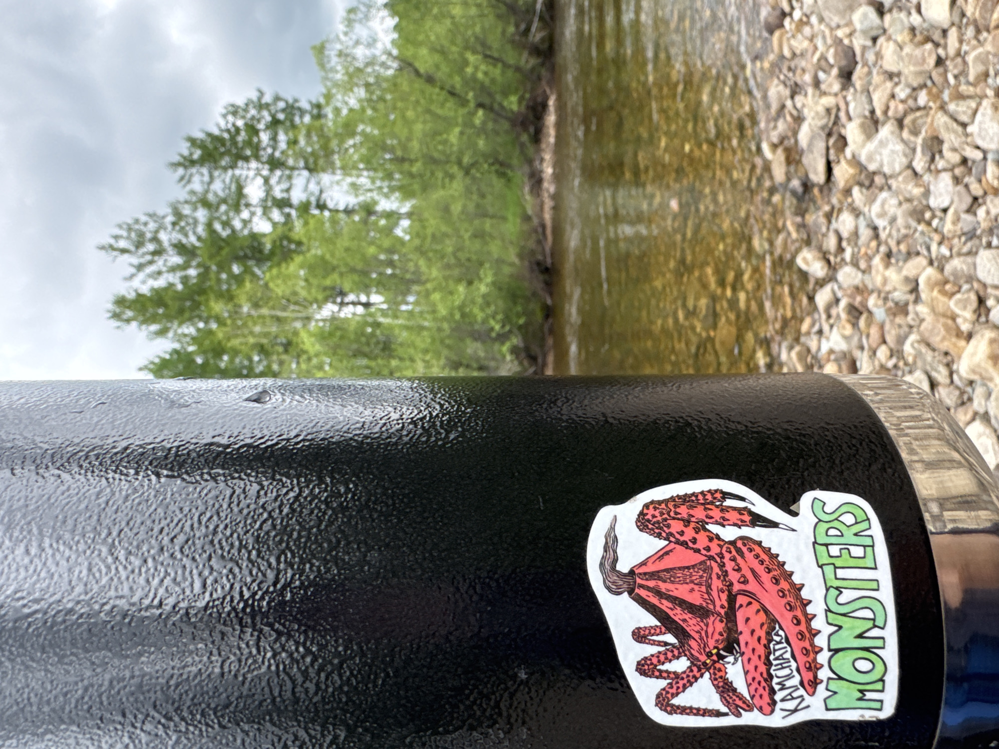 An extreme close-up shot focuses on a black, textured thermos with a sticker of a red crab-like monster and the words "KAMCHATKA MONSTERS" on it. The background is blurred, showing a shallow, clear river with a rocky bed, surrounded by lush green trees and a cloudy sky. The scene appears to be set in a natural environment, possibly near a riverbank. Given the sticker's text, the location might be related to Kamchatka, a peninsula in the Russian Far East. The specific location within the city of Murtygit, Russia, cannot be determined from the image alone.