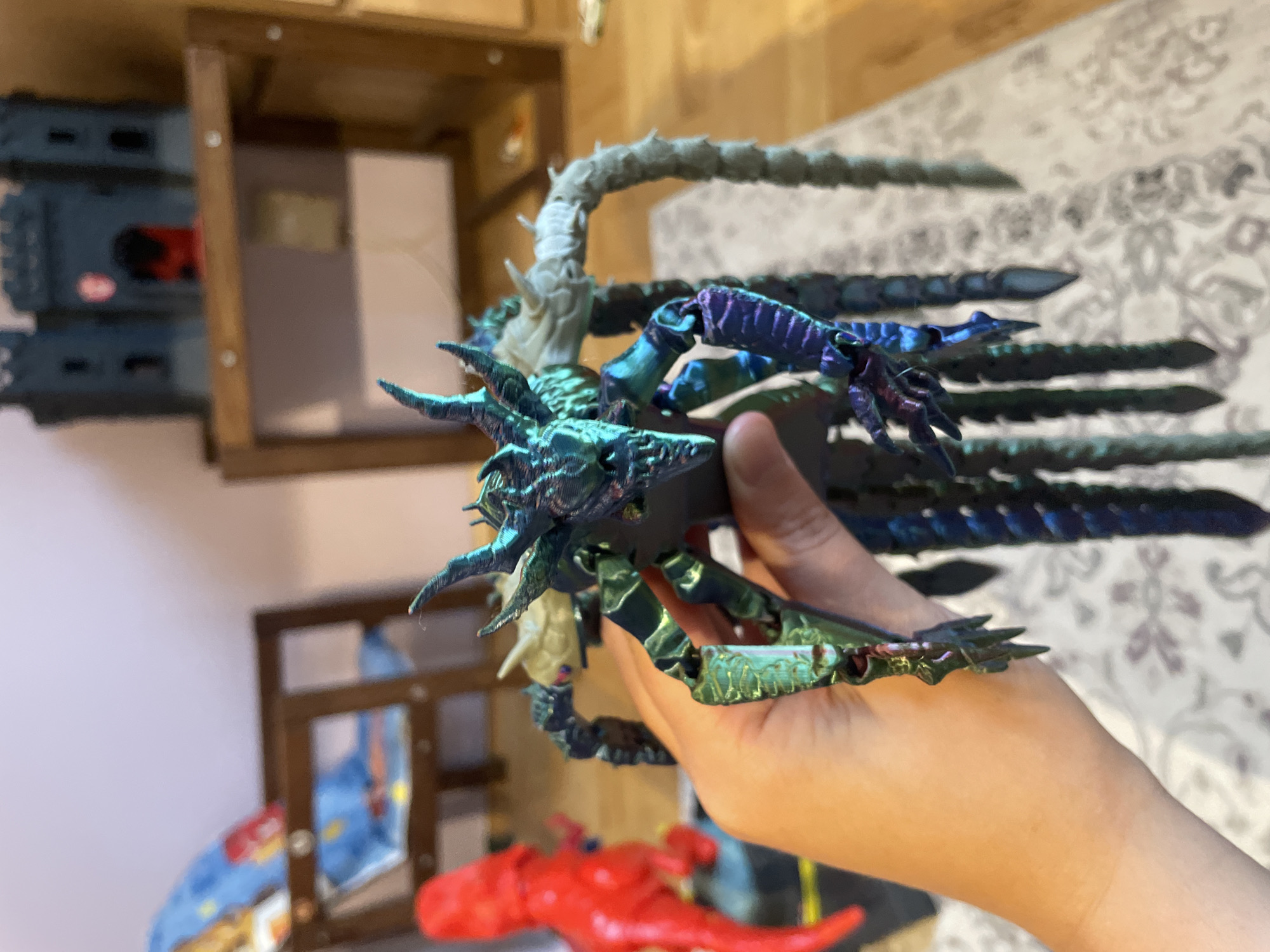 A hand holds a detailed, iridescent toy monster with multiple limbs and a draconic head. The toy has a shimmering blue and green finish, with purple accents on some of its appendages. In the background, out of focus, is a red dinosaur toy, a child's high chair with a blue patterned cushion, and a dark grey toy castle on a shelf. The scene appears to be indoors, possibly in a child's room. The toys are arranged on a patterned rug.