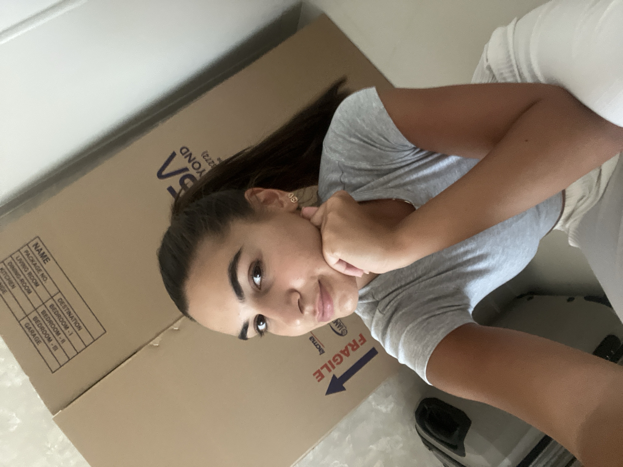 The image shows a young woman posing next to a large cardboard box, likely during a move. The woman has dark hair pulled back into a ponytail and is wearing a gray t-shirt and white bottoms. She is resting her chin on her hand and looking towards the camera. The box has printed information on it, including "NAME", "PACKAGE NO.", "DESTINATION", and lists such as "LIVING ROOM", "DINING ROOM", "BEDROOM-1", "KITCHEN", "BEDROOM-IL", "BEDROOM-", "GARAGE". The word "FRAGILE" is prominently displayed with an arrow. The environment appears to be indoors, possibly inside a room in a house or apartment. The presence of the moving box suggests a transition or change of residence. The image does not include any elements directly indicating a location in Dubai, United Arab Emirates.