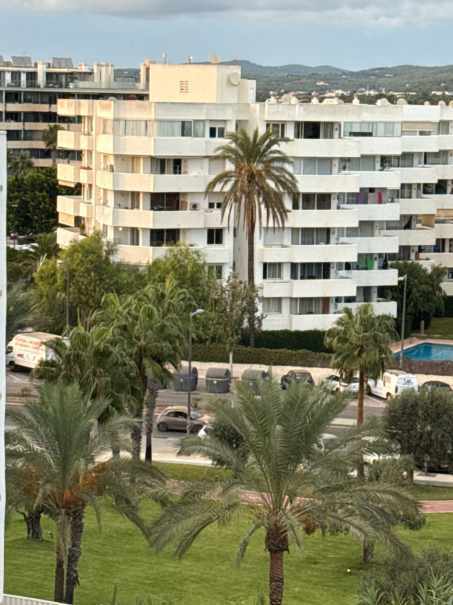 This is a medium-angle, eye-level shot of a residential building in Ibiza, Spain. The building is white, multi-story, and features a stepped architectural design with balconies on each level. Palm trees and other greenery are scattered throughout the scene, with a swimming pool visible in the background. Several cars and a white van are parked on the street in front of the building. The sky is overcast with soft clouds, and the lighting suggests it is either early morning or late afternoon.