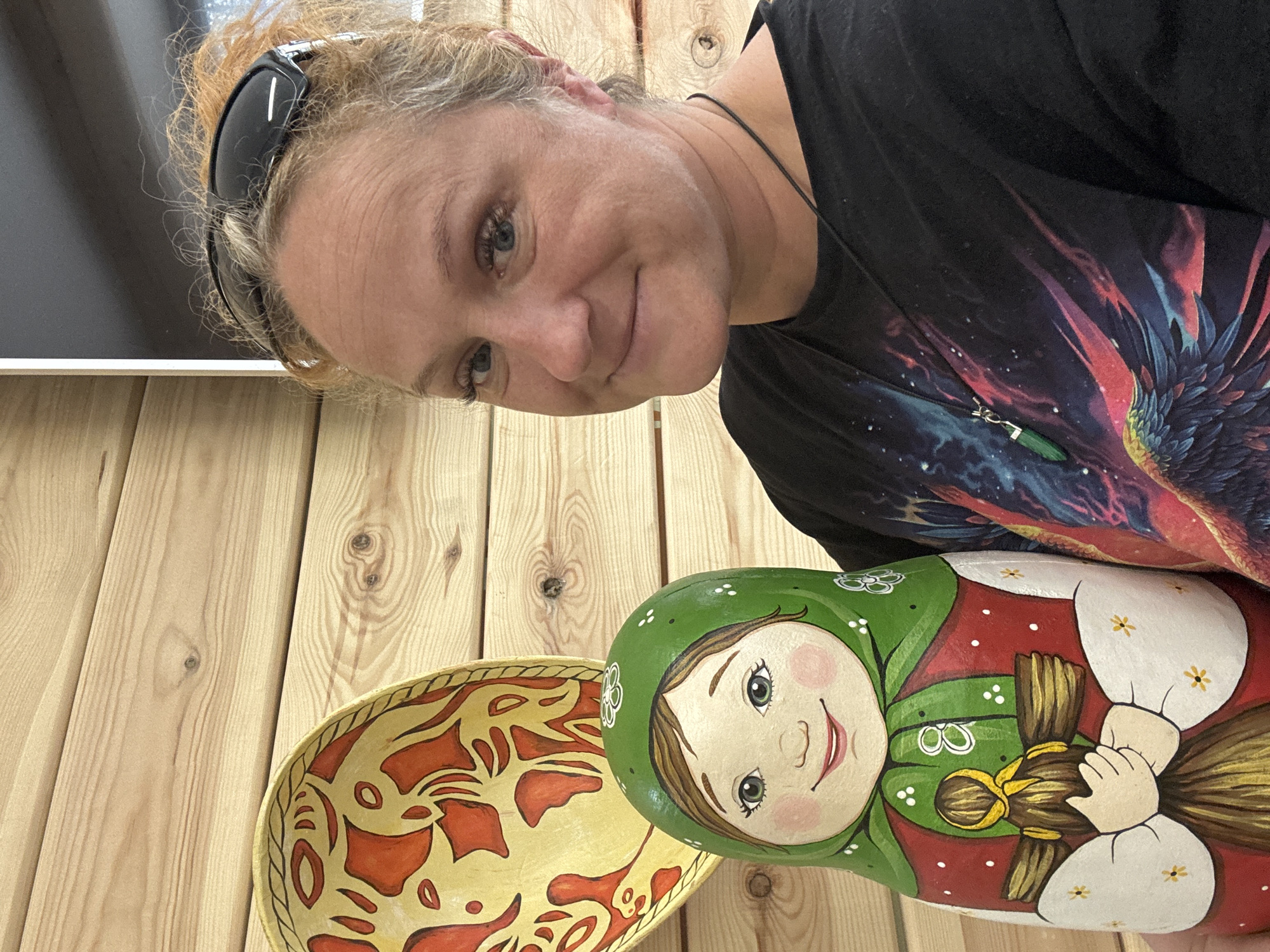 A woman stands inside a building with wooden paneling, posing for a photo. She has blonde hair, sunglasses resting on her head, and wears a black t-shirt with a vibrant print. She is wearing a necklace with a green stone pendant. The woman is holding a painted wooden Matryoshka doll. To the left of the doll is a yellow bowl decorated with red patterns. The precise location in the city of Chaikovsky, Russia, cannot be determined with certainty from the image.