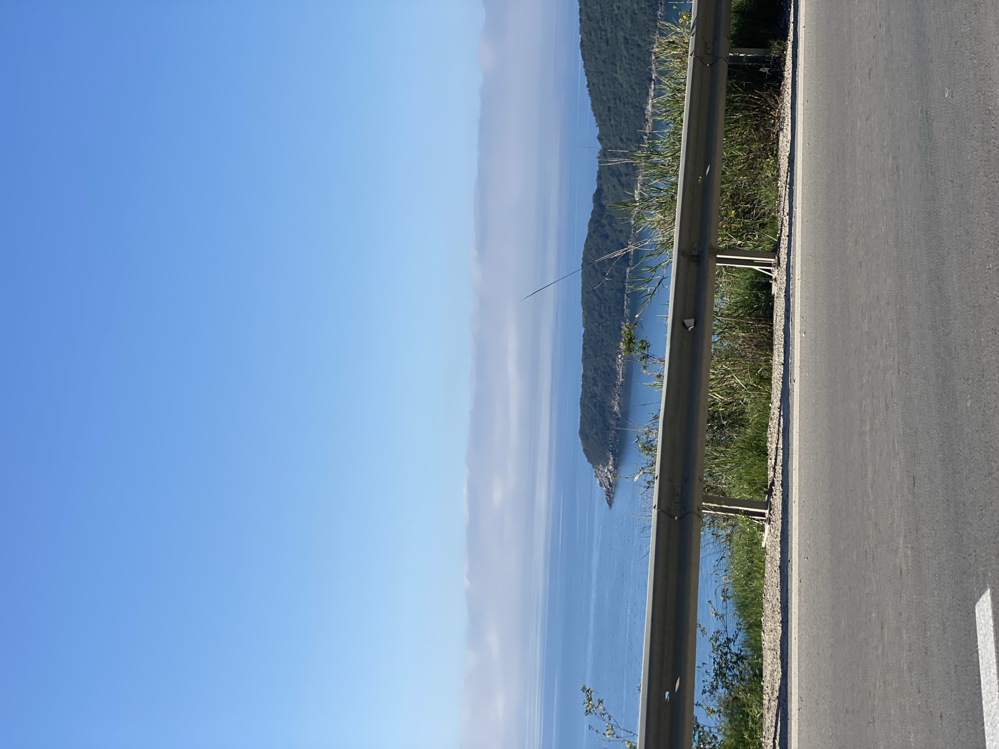 The image depicts a coastal scene near Prijevor, Montenegro. A segment of highway is visible on the right side of the frame, featuring a gray asphalt surface and a roadside guardrail. Beyond the guardrail, a view of the Adriatic Sea stretches towards the horizon. A green, mountainous island punctuates the sea. A cloud layer is between the sea and the island and the distant mountains. The sky is clear blue.