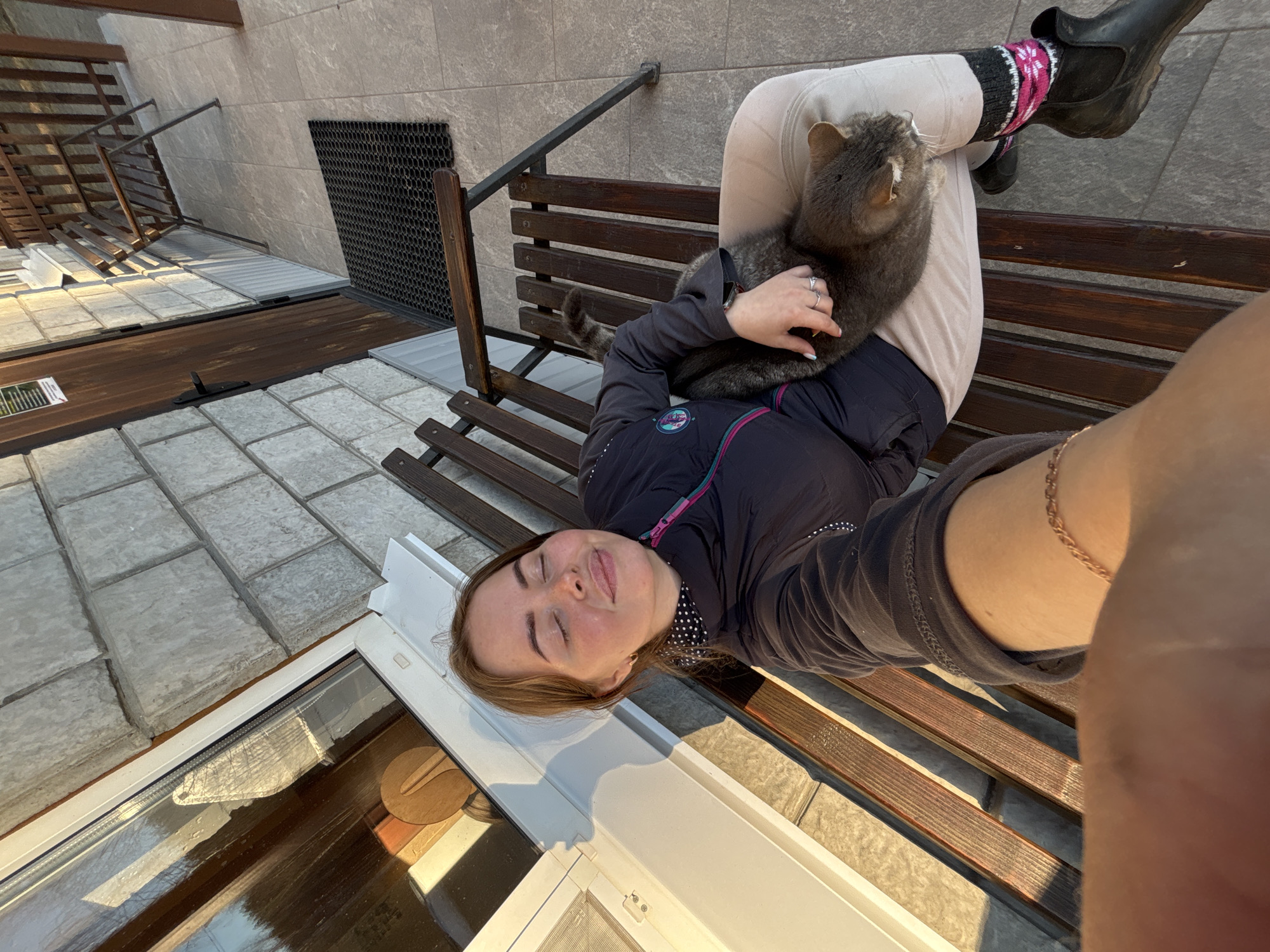 The image captures a woman reclining on a wooden bench, holding a grey cat on her lap. She's wearing light-colored pants, black boots with patterned socks, and a dark jacket. The bench is situated outdoors, adjacent to a building with a grey brick facade. A window and a tiled surface are visible in the foreground, with a railing and another section of wooden structure in the background. There is a gold chain visible on the woman's wrist. The image is taken from a slightly tilted angle. The location appears to be in an urban setting, potentially in Русский Вожой, Russia.