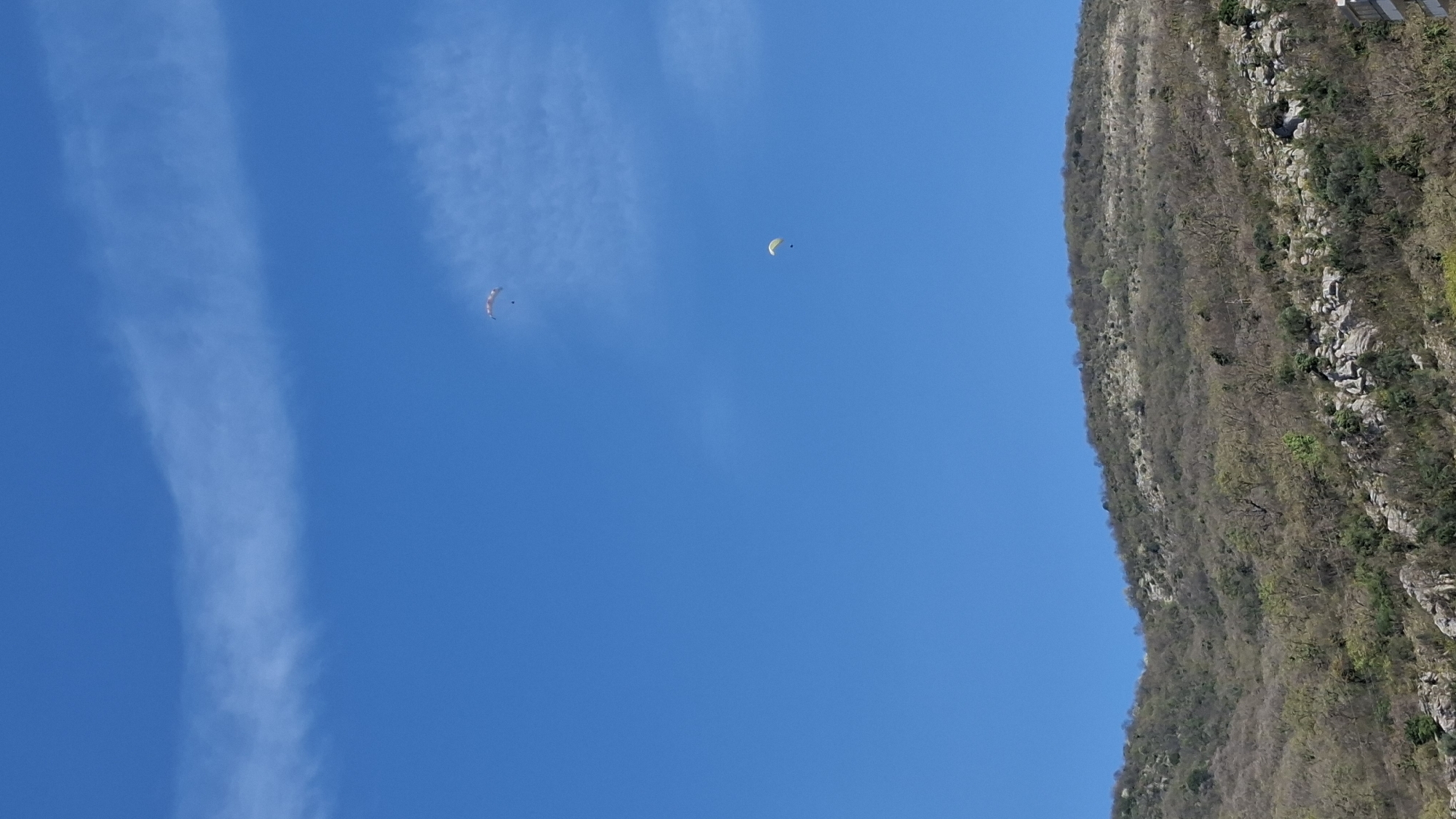 The image depicts a clear, blue sky with two paragliders soaring in the air, one near the center and the other toward the left. Vertical clouds are visible on the left side of the frame. The right side of the image shows a mountain slope covered with sparse vegetation, suggesting a rocky terrain. The scene is likely in or near Budva, Montenegro, due to the mountainous coastal landscape commonly associated with the area. The overall composition is an upward view, emphasizing the height of the paragliders relative to the terrain.