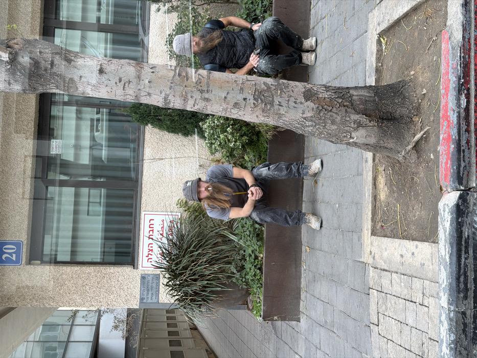 This image depicts an outdoor urban scene in Tel-Aviv, Israel, directly in front of a building marked with the number 20 on a blue plaque. Two men, both with long reddish-brown beards and wearing grey bucket hats, are seated on the concrete ledge of a dark planter box filled with green foliage.
The man on the left wears a grey t-shirt, dark stained pants, and light-colored work boots. A lit cigarette is visible in his mouth, and he holds a yellow object in his clasped hands. The man on the right is partially obscured by a tree trunk in the foreground. He wears a dark vest over a dark t-shirt, dark stained pants, and light-colored work boots, holding a small white object in his right hand. Both men's clothing appears stained, indicative of manual labor.
The building behind them features a textured, light-colored facade with large rectangular windows that reflect the sky and other structures. Below the building number 20, a red sign displays "רחבת הצלה" (Rescue Area) in Hebrew and "FIRE AREA" in English. A smaller white sign with additional Hebrew text is visible beneath the red sign. In the immediate foreground, a tree with light, peeling bark rises from a square patch of bare soil on the grey-tiled sidewalk. The sidewalk is bordered by a curb painted black with sections of red and white.