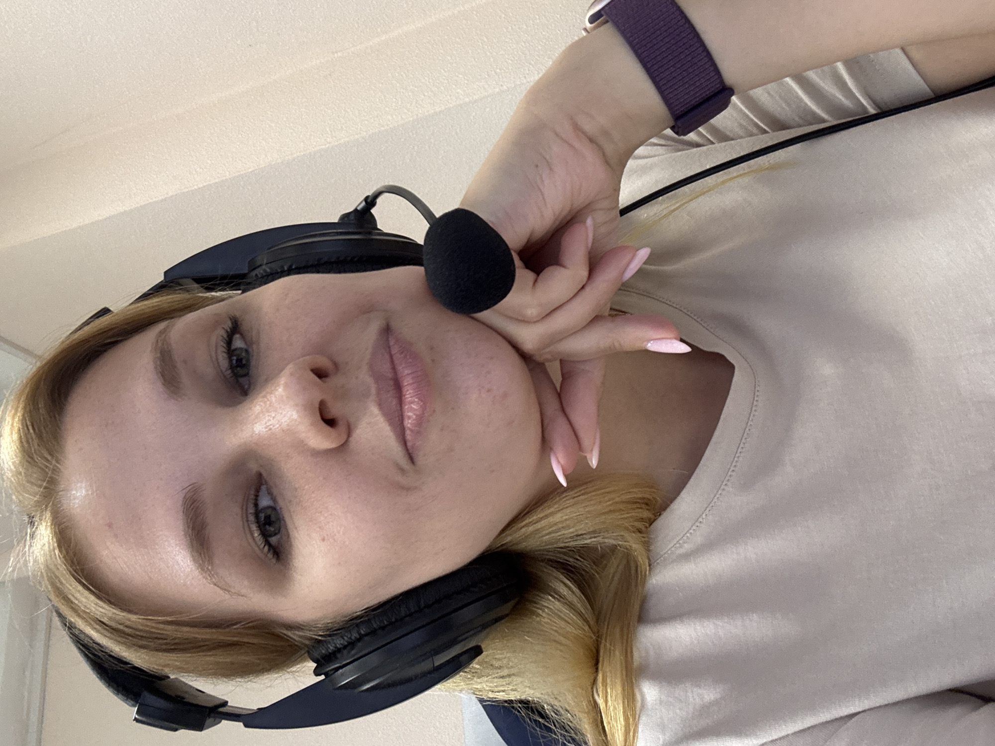 A young woman with blonde hair, wearing a light-colored t-shirt and headphones with a microphone, is captured in a close-up, selfie-style shot. Her left hand rests on her cheek, with manicured pink nails visible. She is looking towards the camera with a neutral expression. The background appears to be an indoor setting with a textured ceiling. A purple smartwatch is visible on her wrist. The image is oriented vertically, as if taken with a phone. There are no visible text elements. The context provided indicates the location is Заволжье, Russia.