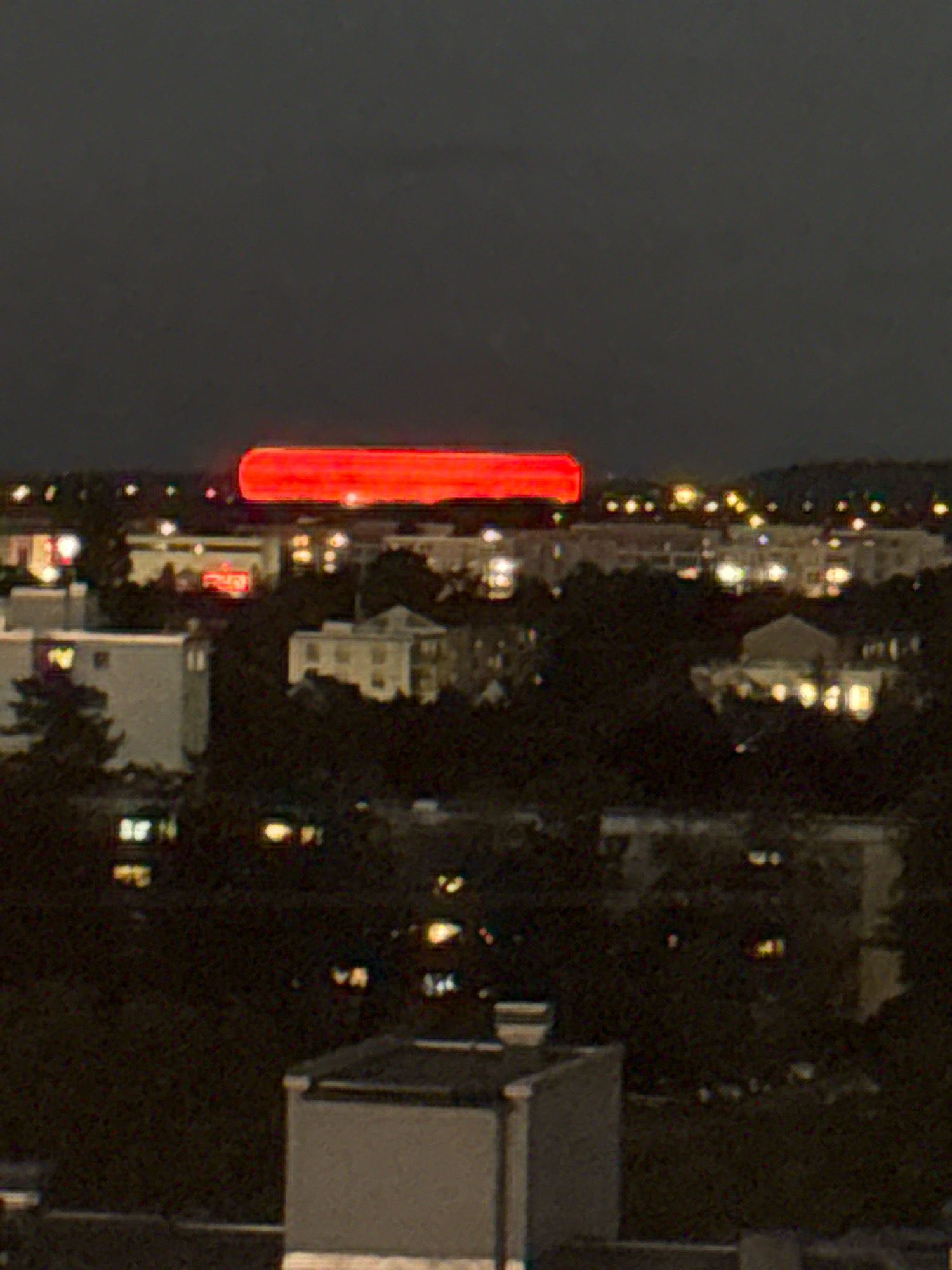 The image shows a blurred nighttime cityscape, likely from a high vantage point. A bright, horizontal red light dominates the upper center of the frame, appearing to be a large illuminated sign or structure. Below this, various buildings with illuminated windows are visible, interspersed with dark trees. The overall impression is of a suburban or outer-city area at night. Without further context or identifiable landmarks, pinpointing the exact location within Munich, Germany, is not possible from this image alone.
