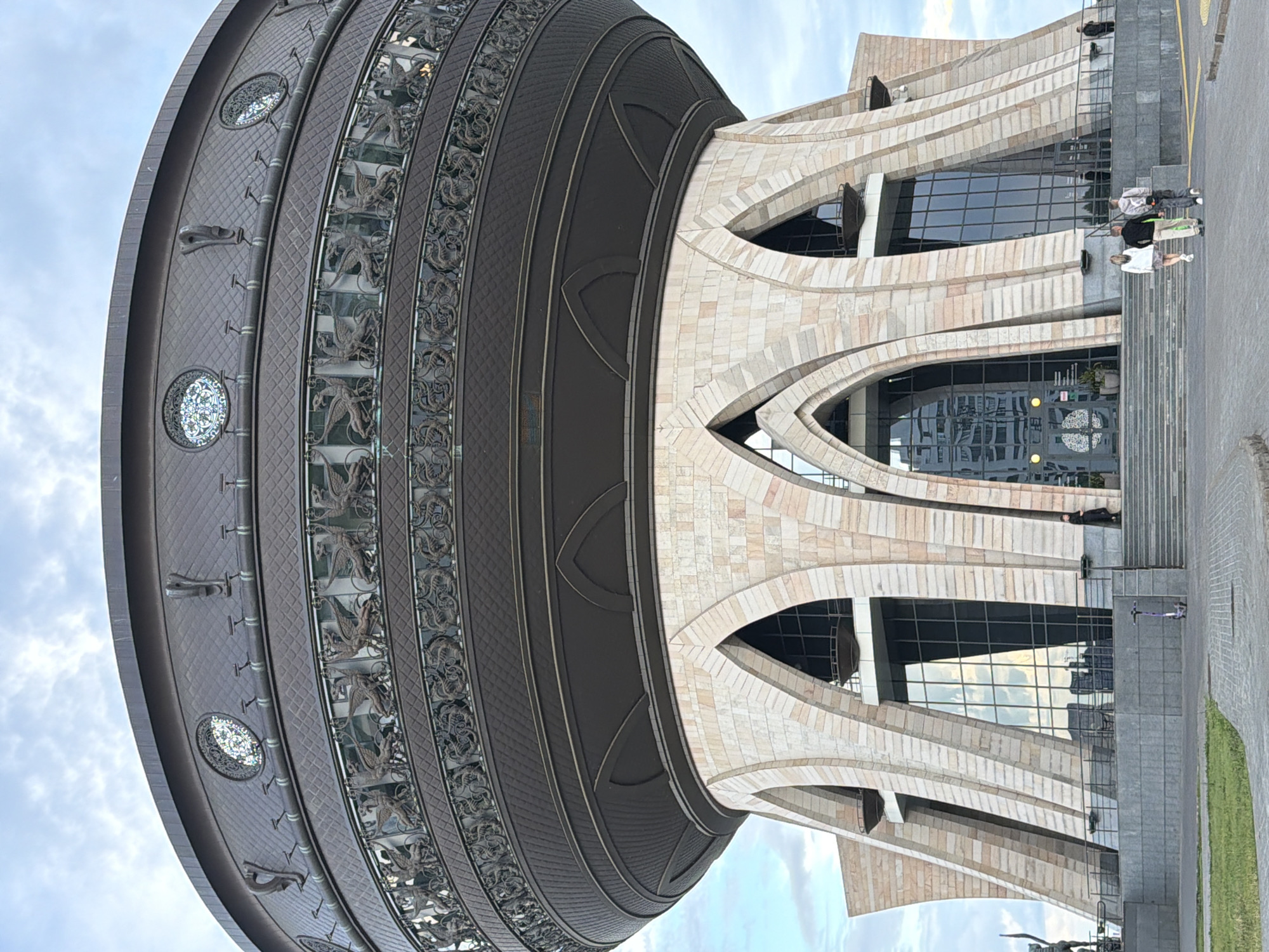The image shows a low-angle, close-up view of the exterior of the National Library of Tatarstan in Kazan, Russia. The building's unique architecture is prominently displayed, with a large, dark, cauldron-shaped upper section featuring ornate metalwork and circular decorative elements. This upper section rests on a base of pale stone blocks, arranged in a series of arched supports that create large openings for windows and entrances. The stone facade has a textured, layered appearance. In front of the building, there are several people walking and standing. Two individuals are walking away from the camera on a paved walkway leading towards the building's entrance. Another person is visible standing on the steps to the right, and a figure is standing near a statue on the far left. A purple scooter is parked on the lower level. The sky above is cloudy, with patches of lighter sky visible. The overall scene captures a portion of the striking and unconventional architecture of the library against a natural backdrop.