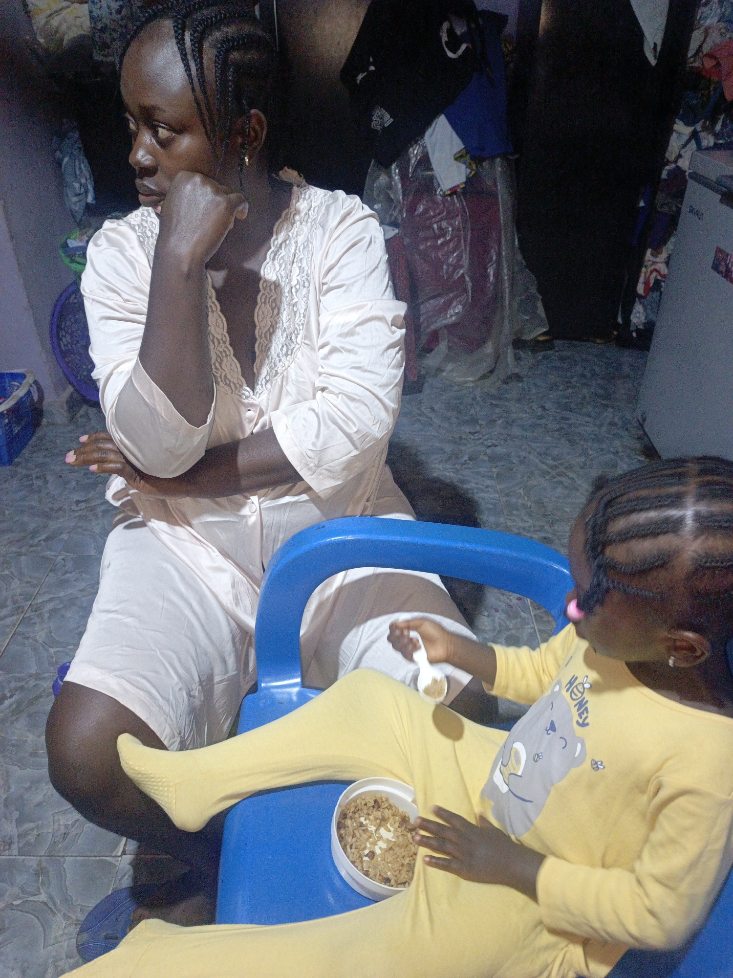In an indoor setting, an adult woman with braided hair, wearing a pale pink nightgown, is seated with her right arm resting on her chin and her left arm bent. Beside her, a child in a yellow pajama set, also with braided hair, is seated in a blue chair, holding a white spoon with food. A white bowl of food is placed in front of the child. The floor is tiled, and in the background, various objects and clothing items are visible, including a blue laundry basket and a light-colored appliance. The lighting is dim, creating shadows throughout the scene.