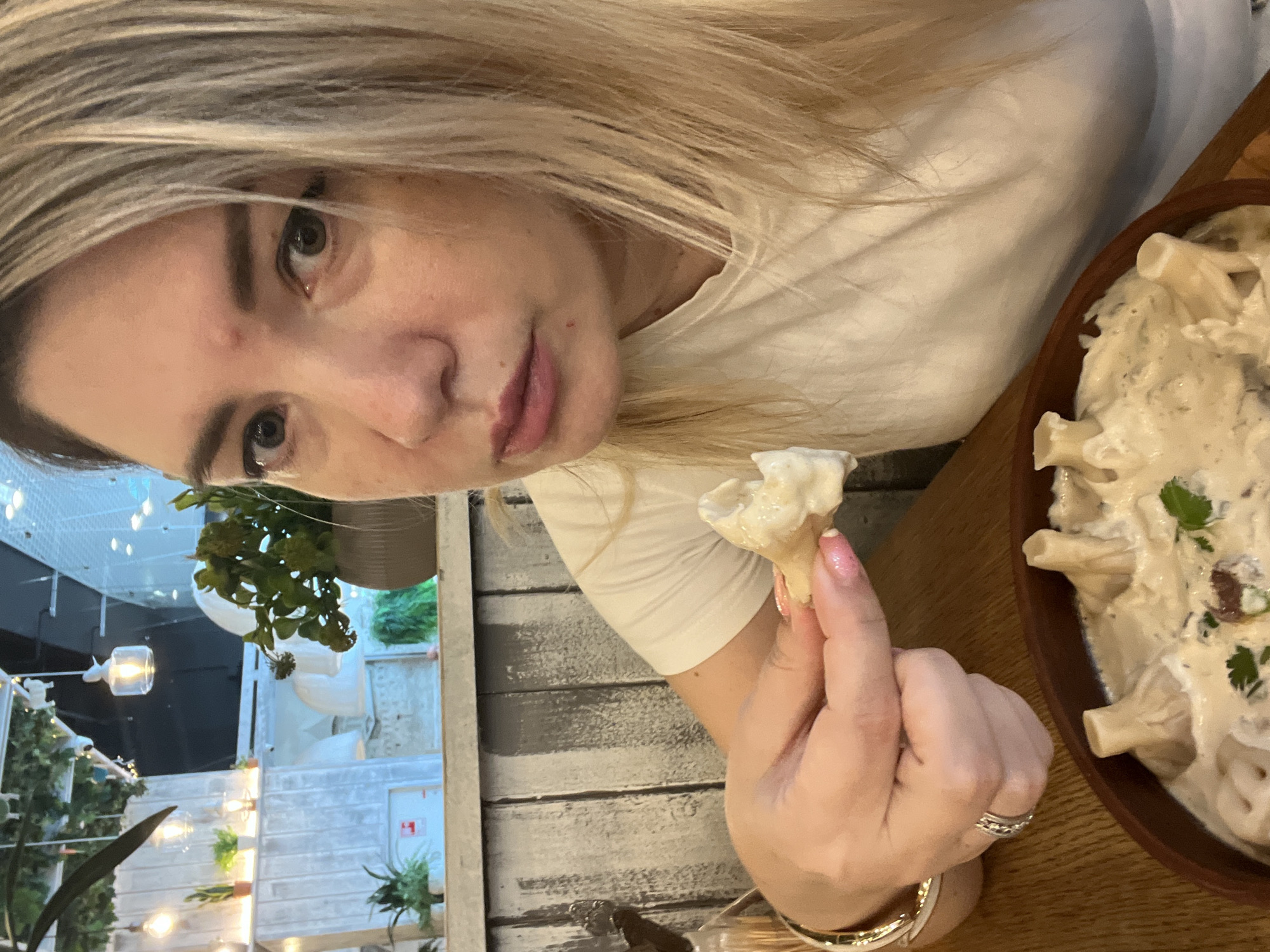 A woman with blonde hair and light skin is holding a piece of food in her right hand, bringing it towards her mouth. She is wearing a white t-shirt and a ring on her ring finger. In front of her, on a wooden table, is a bowl of food that appears to be pasta with a creamy sauce. The background shows a restaurant interior with wooden paneling, plants, and lighting fixtures. The environment suggests a casual dining setting in the city of Sochi, Russia.