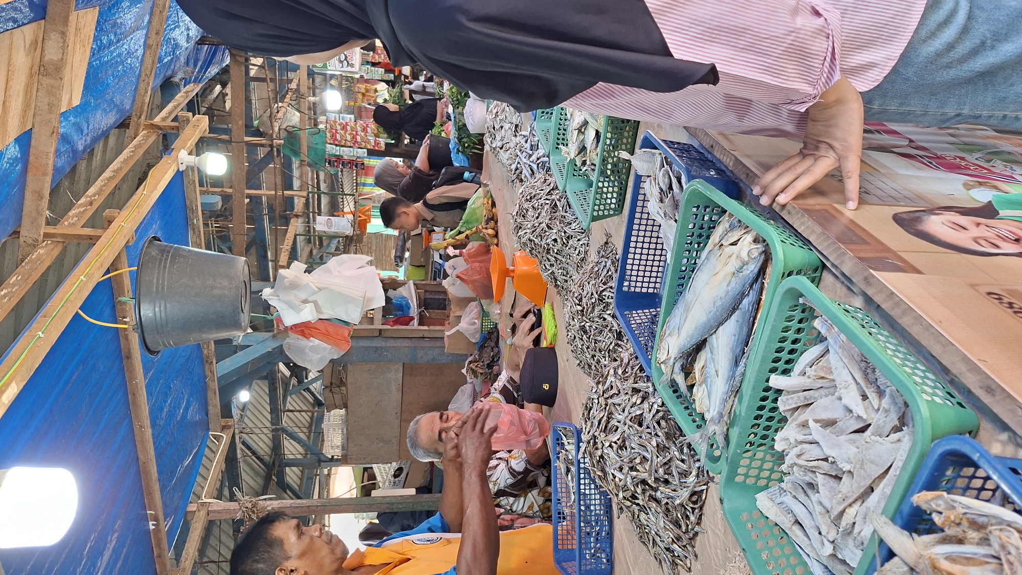 This is a photograph taken at an outdoor market in Dayahbaro, Indonesia. The scene is busy with people and stalls selling goods. In the foreground, a woman wearing a black hijab and a pink striped shirt stands facing away from the camera, looking at a market stall. Her hands are resting on the edge of the stall. The stall is laden with various types of dried fish arranged in green and blue plastic baskets. Two large, whole dried fish are visible in a green basket, and a pile of smaller dried fish is in a blue basket. Behind the woman, a man wearing an orange vest and blue shirt appears to be selling to an elderly man. The elderly man, wearing a patterned shirt and a black cap, is holding a pink plastic bag. Next to him is an orange scale. In the background, other people, including children, are visible browsing stalls. The market is covered by a blue tarp, supported by wooden beams and poles. Hanging from one of the beams is a large black bucket. The overall impression is a bustling marketplace with a focus on fresh and dried seafood.