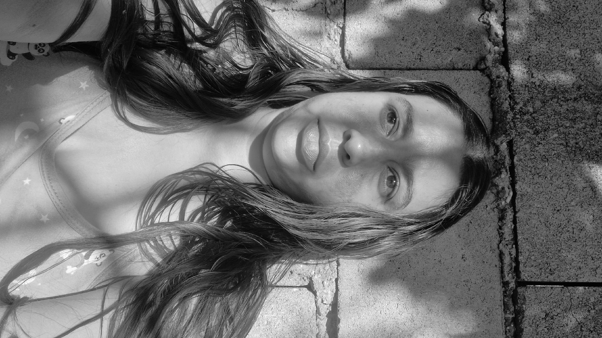 In Vallecito, Venezuela, a close-up, black and white, waist-up photograph captures a young woman posing against a textured, rough-hewn cinder block wall. Sunlight casts dappled shadows across her face and hair, creating a dramatic chiaroscuro effect. She has long, dark, wavy hair that falls around her shoulders, partially obscuring her skin. Her facial expression is directed slightly to her left, with her mouth slightly ajar and her tongue subtly protruding. The wall behind her is weathered and shows signs of age, with visible mortar lines and irregular surfaces. The lighting suggests an outdoor setting during daylight hours, likely in a somewhat rustic or unrefined urban environment characteristic of Vallecito. She wears a patterned garment with faint celestial motifs, including moons and stars, and a small, light-colored charm, possibly a panda, hangs from her neck. The overall impression is informal and playful, a self-portrait taken in natural light.