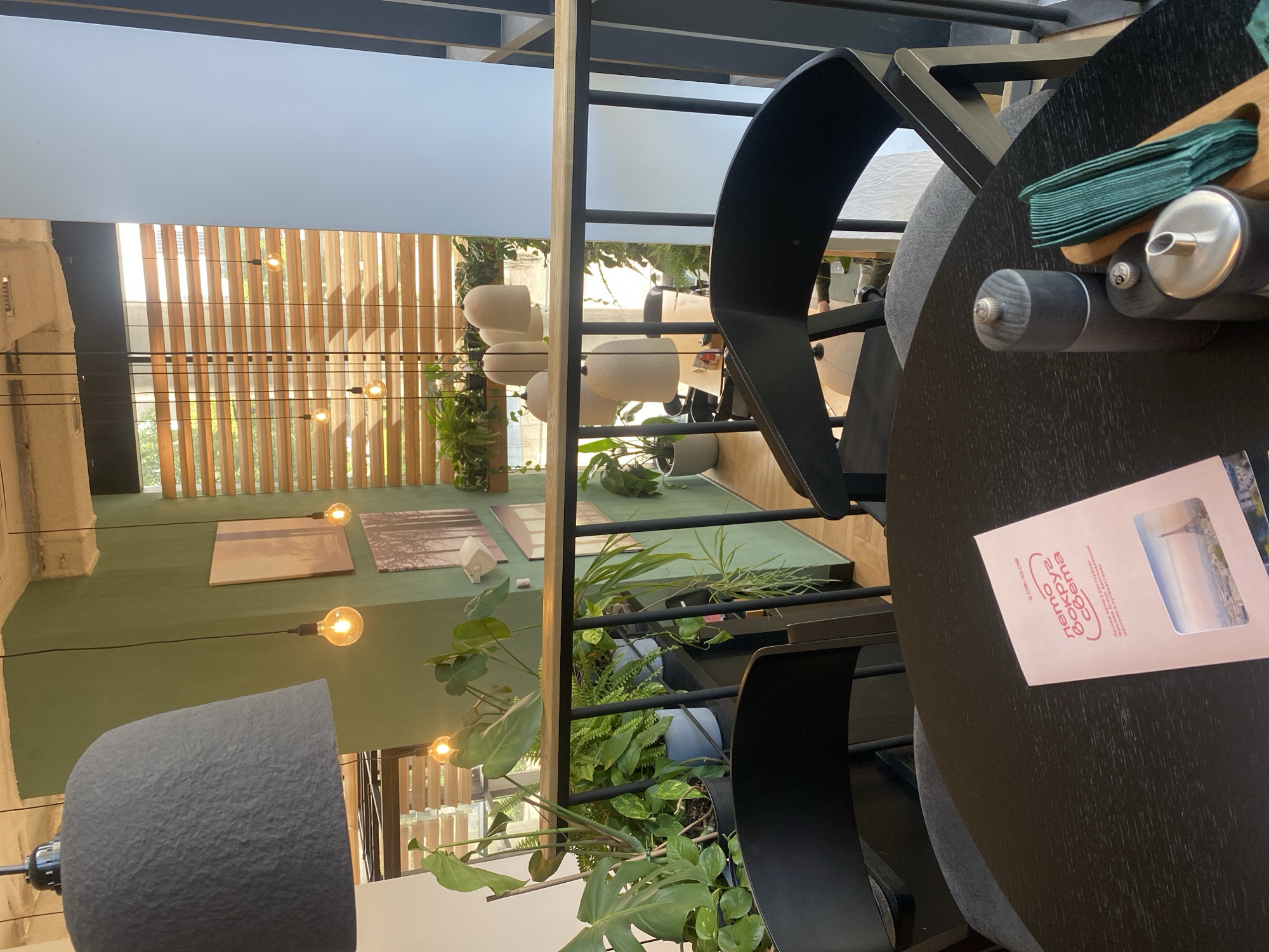A view of a dining area in a cafe with an interior balcony. On the balcony, numerous potted plants are arranged on shelves and a railing separates the balcony from the seating area below. Hanging pendant lights are visible throughout the space. A black circular table is in the foreground, set with a menu, salt and pepper grinders, and a holder with green napkins. Two black chairs are positioned at the table. The cafe has green walls and wooden blinds on the windows. Artwork is displayed on the walls, and the lighting suggests it is daytime.