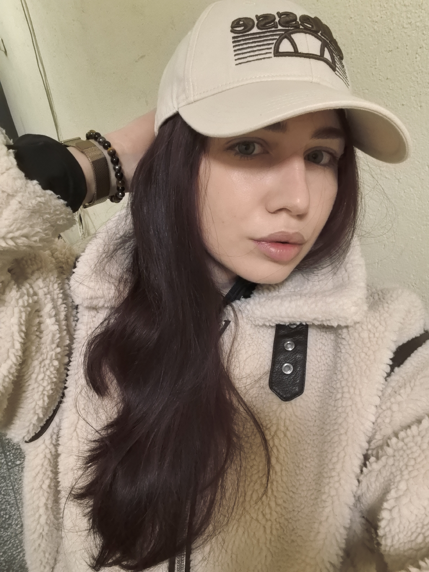 A young woman poses indoors, likely in an apartment. She is wearing a light beige baseball cap with a dark logo, a textured cream-colored sherpa-style jacket with leather detailing on the shoulders and closure, a gold watch, and a dark beaded bracelet. She has long, dark hair. The background is a plain, light-colored wall. No information about location in Stara Zagora, Bulgaria can be ascertained from the image.