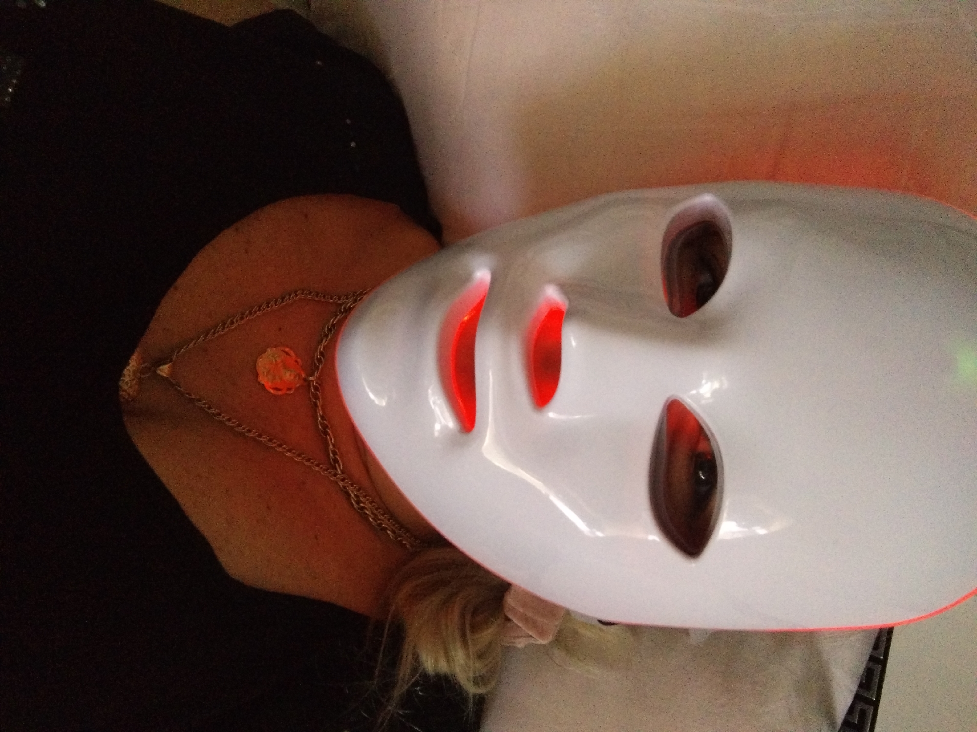 A person is wearing a white LED facial mask with red light emissions visible through the eye and mouth openings. The mask covers the entire face. The person is also wearing a black top and a gold chain necklace with a circular pendant. Their blonde hair is visible on the right side of the image. The background is blurred and appears to be a soft, textured surface. There is no discernible information to suggest a location within Stara Zagora, Bulgaria.