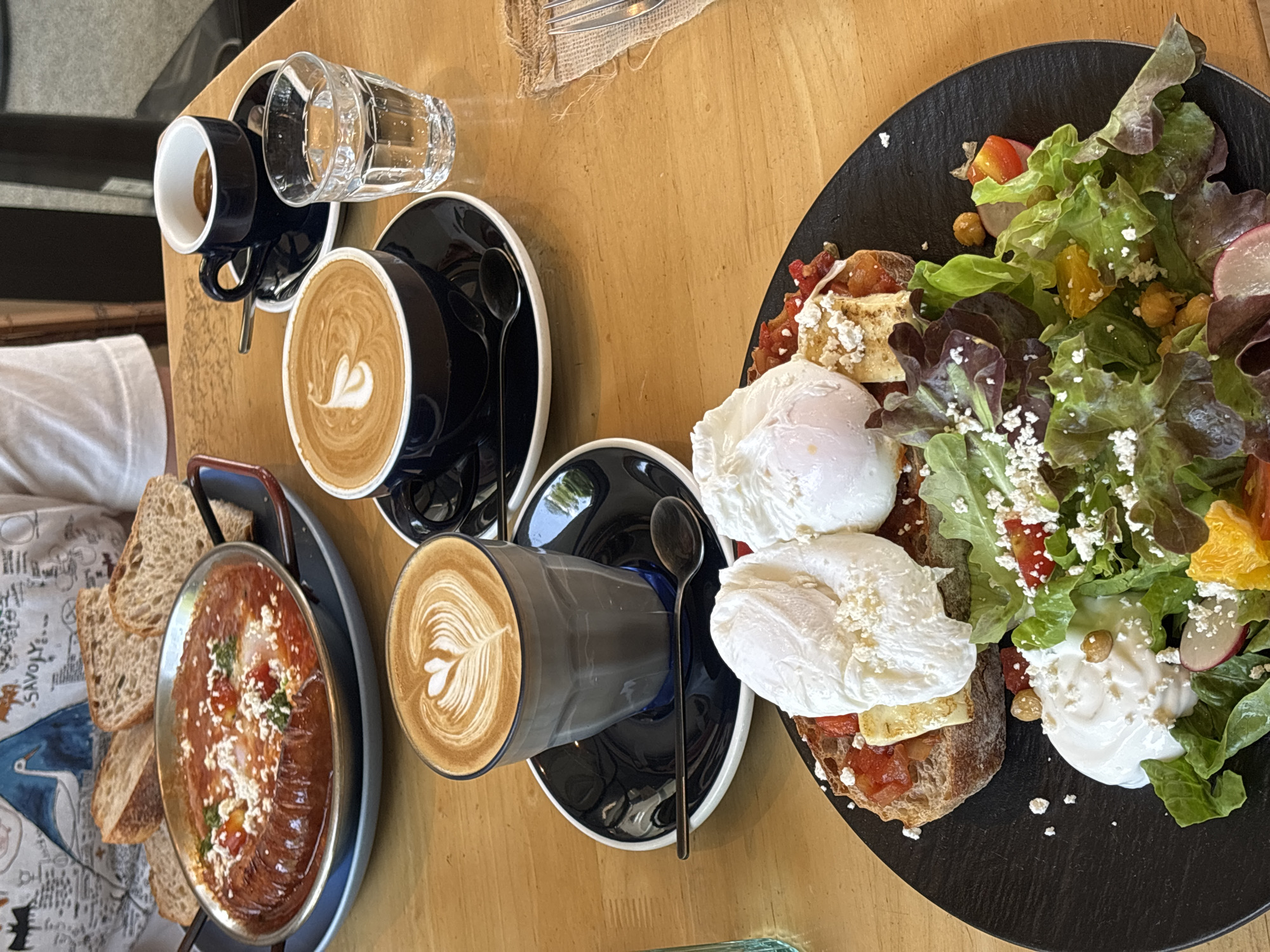 An overhead shot depicts a table laden with brunch items, likely in a cafe in Choeng Thale, Thailand. From left to right: a tray with a dish of shakshuka and sliced bread sits near a person's arm. Next to it, a small cup of espresso, a glass of water, and two cups of coffee are placed on matching saucers. On the far right is a black plate with toast topped with poached eggs, a salad of lettuce, radishes, chickpeas, and diced oranges with feta cheese.