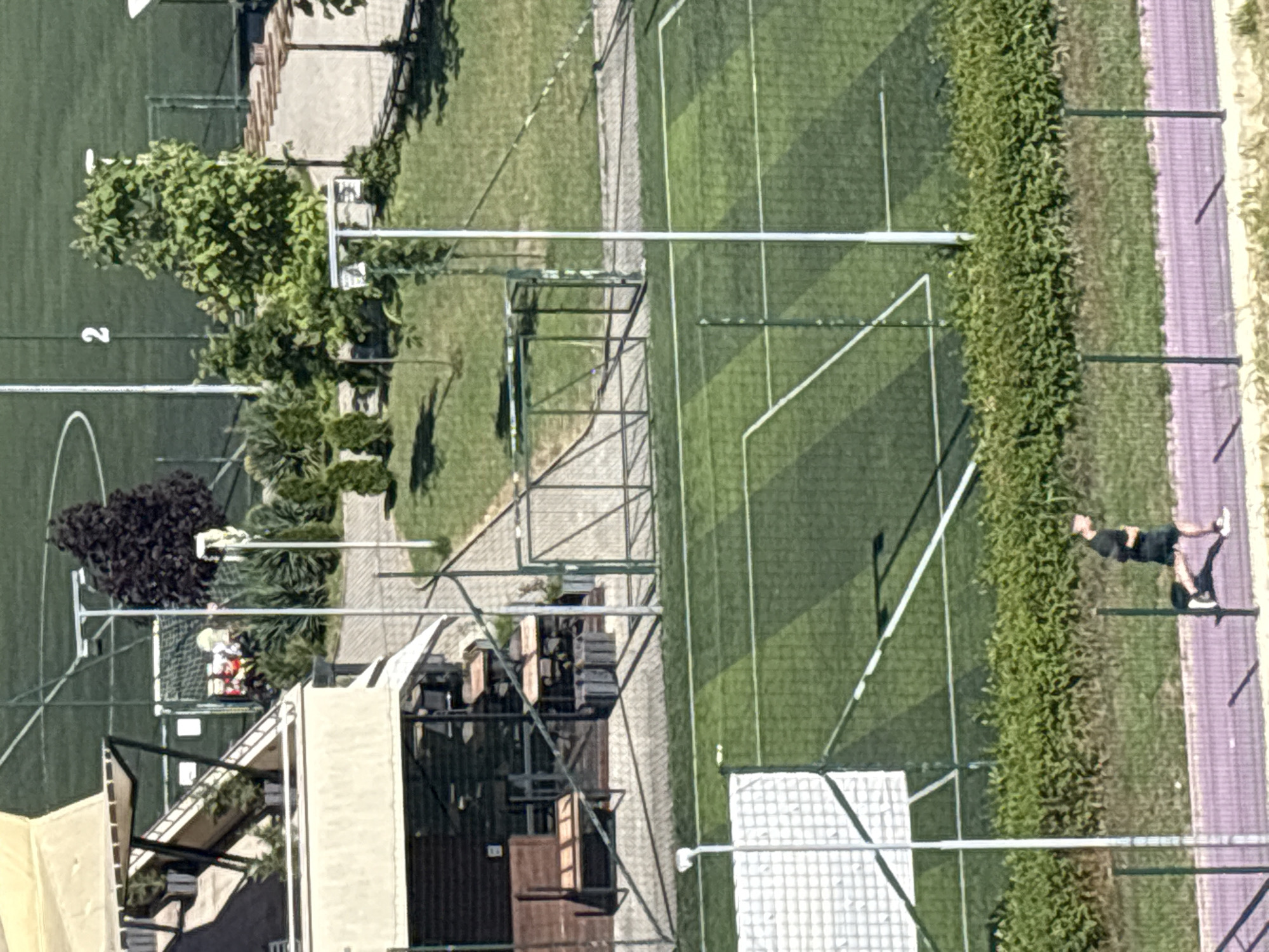 This is an aerial view of an outdoor sports facility with artificial turf fields. The scene appears to be a sports complex or park. In the foreground, there is a building with a light-colored awning and a dark-colored roof. Adjacent to the building is an outdoor seating area with tables and chairs, partially covered by a metal structure.
A person wearing a dark t-shirt and shorts, along with sneakers, is visible on the right side of the image, appearing to walk or jog along a pathway next to a green hedge. The sports field itself is marked with white lines, suggesting it is set up for a sport like soccer or football. There are also some metal structures, possibly goals or fences, present on the field. In the background, there are trees and more green spaces.
The lighting suggests it is daytime, likely sunny given the clear shadows cast by the structures and the vibrant colors of the scene. There are no explicit time-of-day indicators or weather cues beyond the sunshine. No visible text is present in the image. The overall impression is of a well-maintained recreational area.