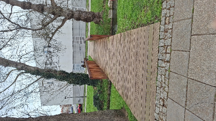 A wooden boardwalk extends into a green space, bordered by grass and low shrubs. A building with multiple windows is seen behind trees with bare branches. Part of a red car is visible near the building. The boardwalk connects to a stone path, composed of various stone sizes and colors, on the right side.