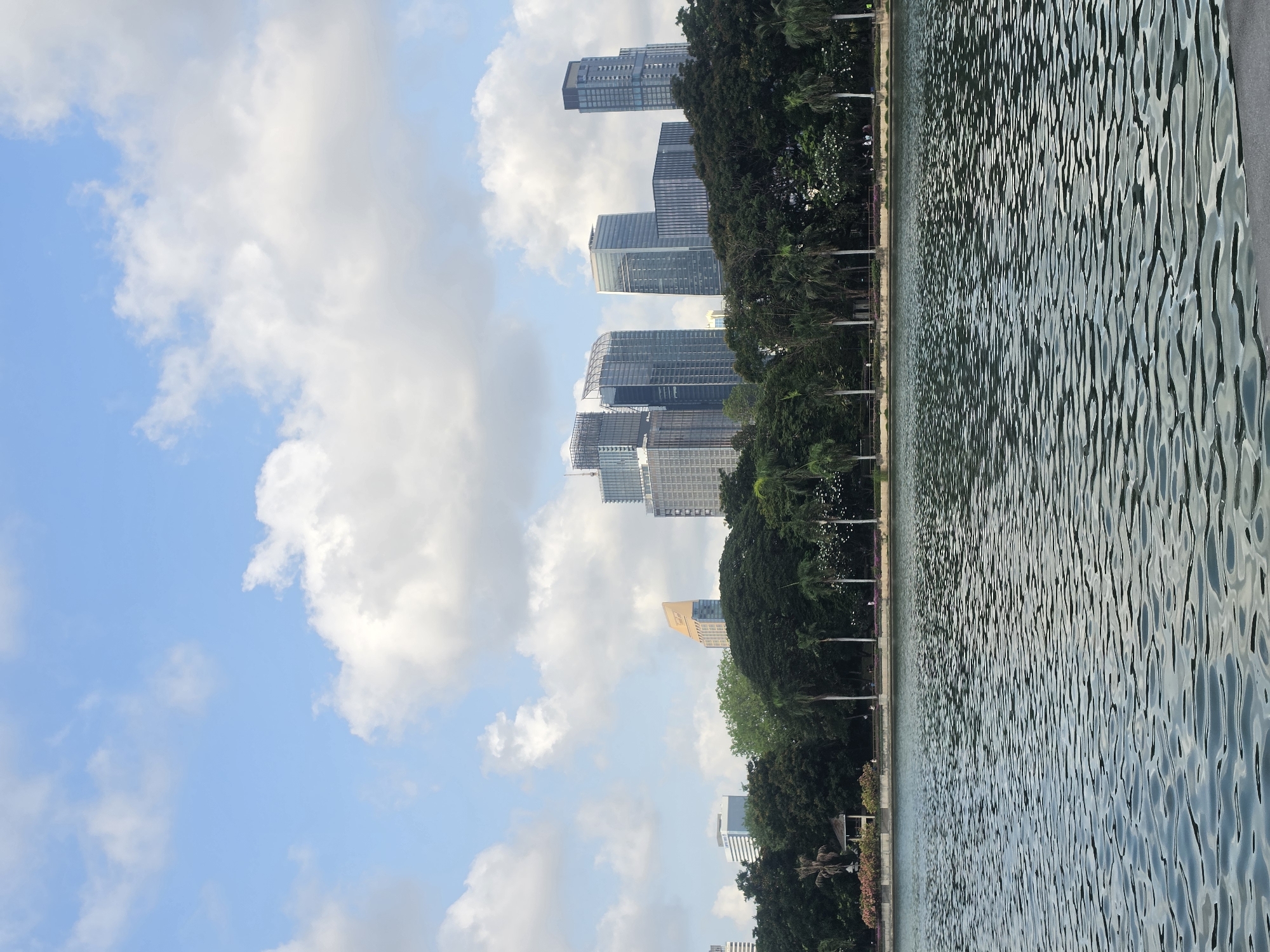 The image depicts a water body in Bangkok, Thailand, with skyscrapers in the background. Lush green trees line the edge of the water. The sky is blue with scattered white clouds.