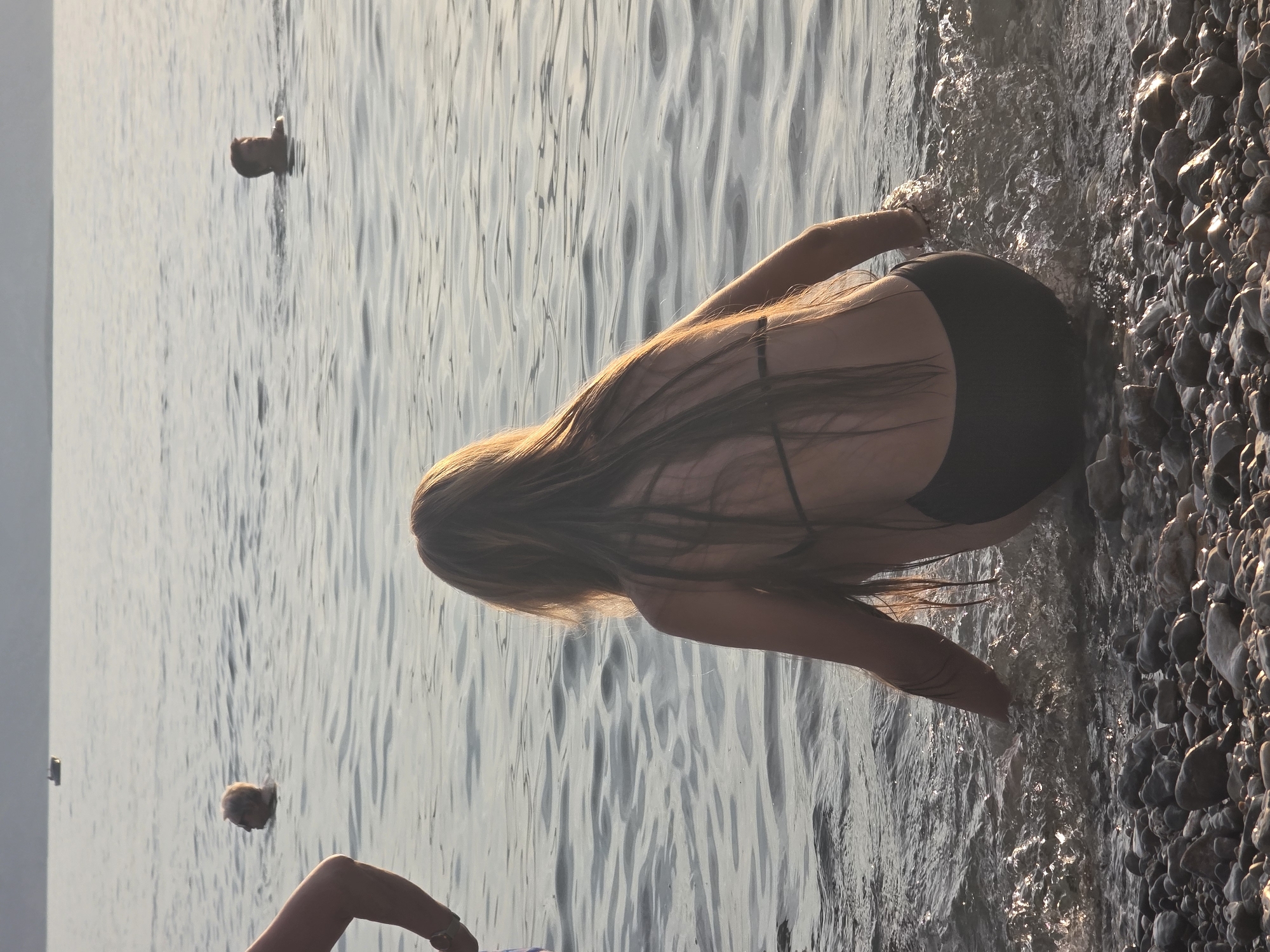 A medium-shot, eye-level, outdoor photograph captures a group of people in a body of water near a pebble beach. In the foreground, a young woman with long, light brown hair is seated at the water's edge, facing away from the camera and towards the water. She is wearing a black bikini. Her left arm is extended into the water, and ripples are visible around her hand. The beach is covered in smooth, grey and white pebbles. In the mid-ground, a man with short, grey hair is swimming in the water, his head and shoulders visible. Further in the distance, another man with dark hair is also swimming. A small boat is visible on the horizon. The water is calm with gentle waves, and the lighting suggests it is late afternoon or early evening. The background is a hazy silhouette of a distant shore.
