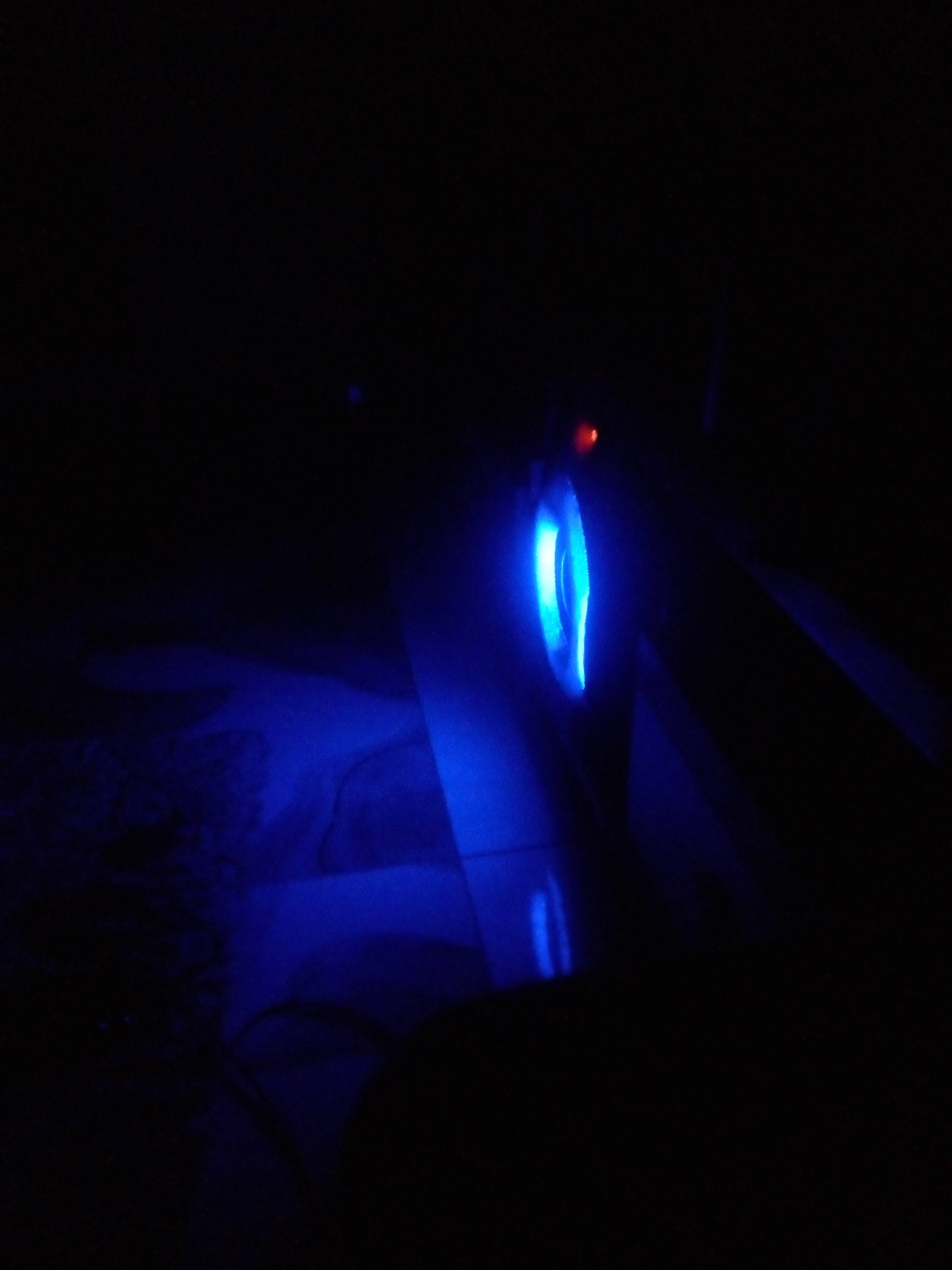In a dimly lit environment, likely at night, a blue LED light emanates from a circular object, possibly a computer mouse or a similar electronic device. The blue light reflects and casts shadows on the surrounding surfaces, creating an abstract and atmospheric scene. A small, red light is also visible near the blue light source. The background is largely dark and indistinct, suggesting an indoor setting with minimal ambient light. The scene appears to be devoid of people, with the focus solely on the illuminated object and its effect on the environment. The exact location in Nyanya, Nigeria, is not discernible from the visual information provided.
