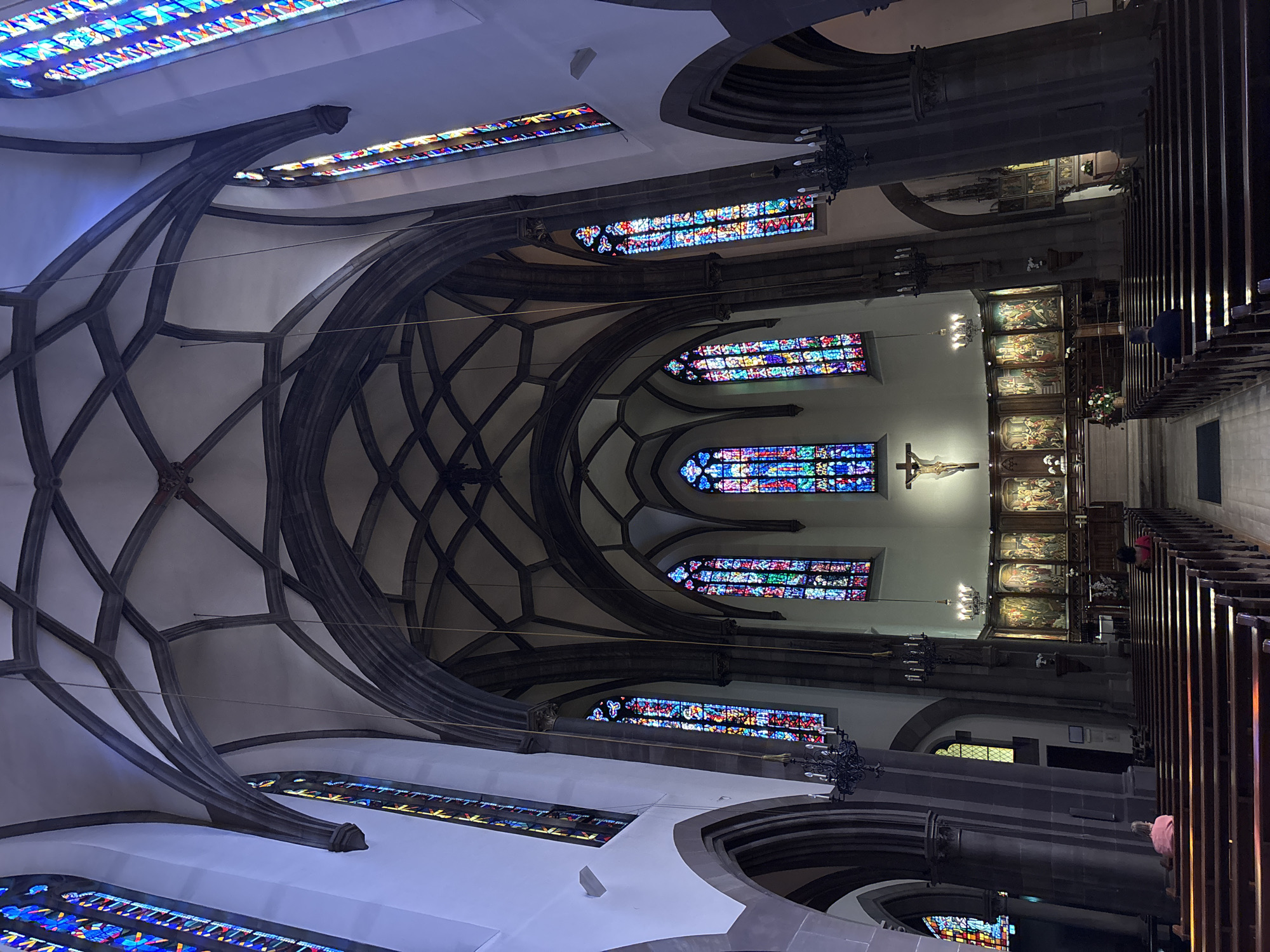 This is an interior shot of a grand Gothic cathedral, likely Saint-Pierre-le-Jeune Protestant Church in Strasbourg, France. The image captures the vast nave with its high, arched ceilings and intricate stone vaulting. Sunlight streams through tall, vibrantly colored stained-glass windows lining the sides of the nave, casting colorful patterns on the pale walls.
At the far end of the nave, a large crucifix hangs above a series of ornate altarpieces depicting religious scenes. The main aisle is lined with rows of dark wooden pews. Several individuals are seated on the pews, facing towards the altar, suggesting a service or quiet contemplation. One person in the foreground is seated in the left aisle, facing away from the camera, wearing a pink shirt. Another person is seated in the right aisle, also facing away, wearing a dark blue jacket.
The environment is one of solemnity and grandeur, typical of a historic religious institution. The architecture emphasizes height and light, with dark, imposing columns supporting the soaring arches. Chandeliers hang at intervals, adding to the ambient lighting. The floor appears to be a light-colored stone or tile. The overall impression is one of immense space and detailed artistry.