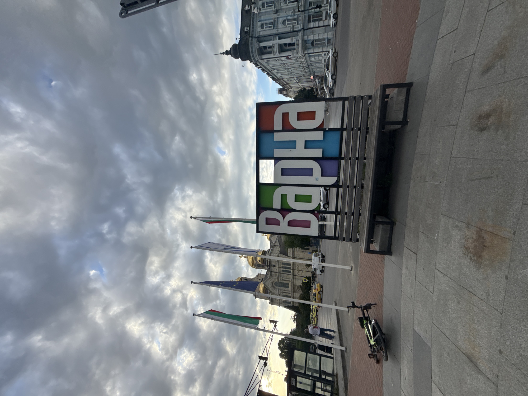 The image shows a street scene in Varna, Bulgaria. A bench with the word "Варна" (Varna) displayed in colorful letters stands on a gray-paved area. Behind the bench are three Bulgarian flags, and one European Union flag. In the background, the Cathedral of the Assumption of the Virgin is visible. A light-colored building is also visible. A bicycle is parked. A person is walking nearby. The sky is cloudy.