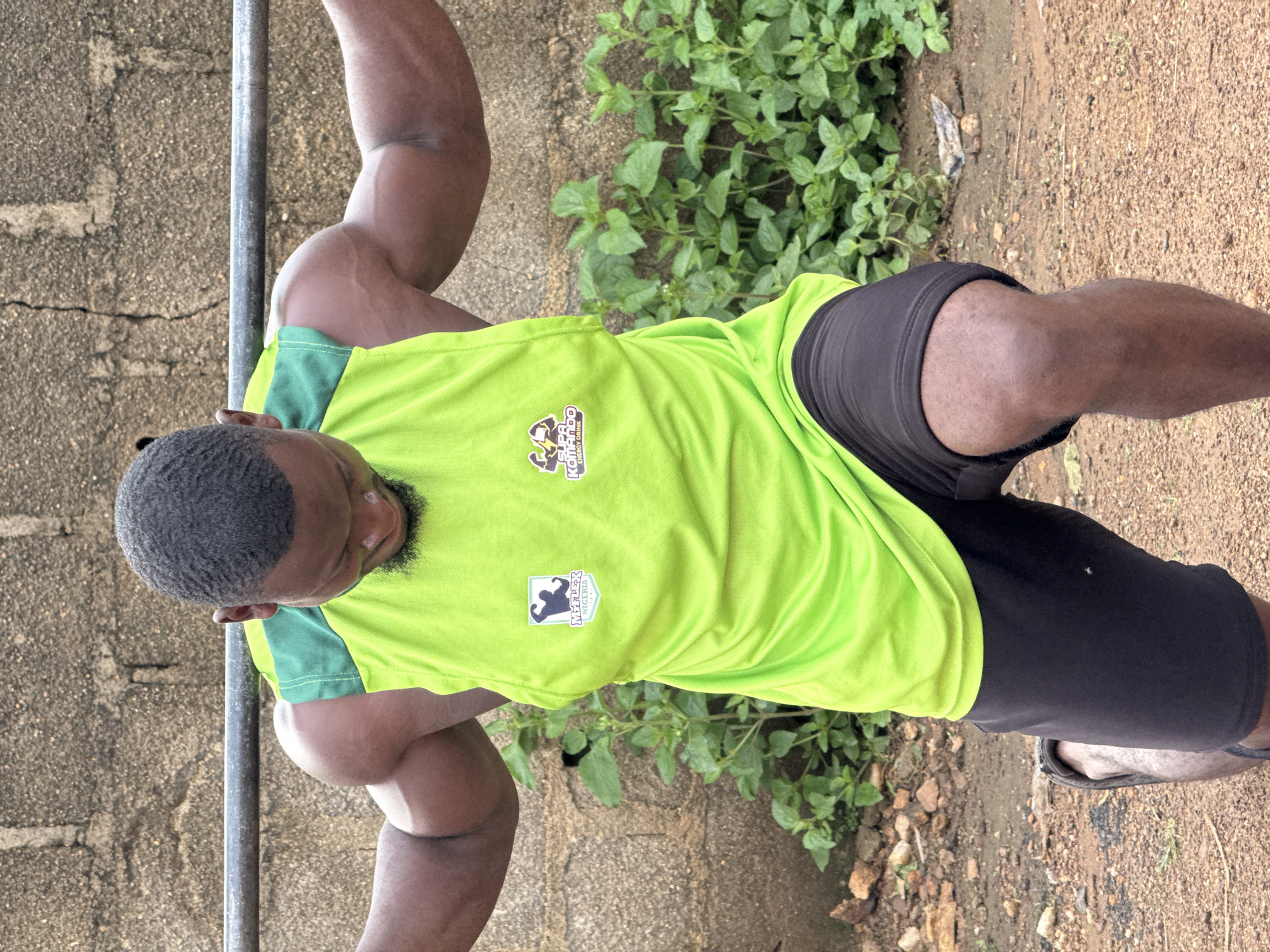 In the city of Akwanga, Nigeria, a Black man is captured in a medium shot from the chest down, performing a lunge with a barbell across his upper back. He is wearing a bright green sleeveless athletic shirt with green accents and black shorts. His muscular physique is evident, particularly his toned arms and legs. The background consists of a rough, grey brick wall and a patch of lush green foliage, suggesting an outdoor training area. The ground is a mix of dirt and small stones. The lighting is natural, indicating it is daytime. A logo with the words "SUPA KOMANDO ENERGY DRINK" is visible on the right side of his shirt, and another logo with "MYFLEX NIGERIA" is on the left.
