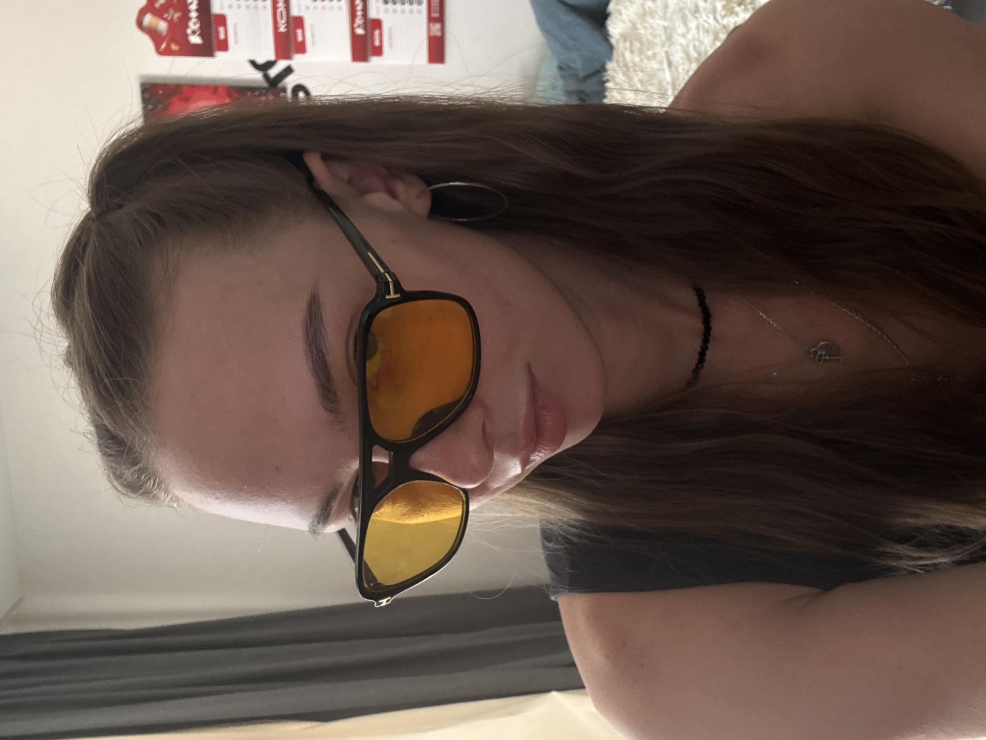A medium close-up shot shows a young woman wearing dark-rimmed aviator sunglasses with orange lenses. She has her hair pulled back in a ponytail and is looking downwards. She is wearing a dark-colored tank top and a beaded choker necklace with a silver pendant. She also has a thin silver chain necklace with a key-shaped pendant. There are hoop earrings in her ears. The background shows a light-colored wall with some posters and a curtain on the left.