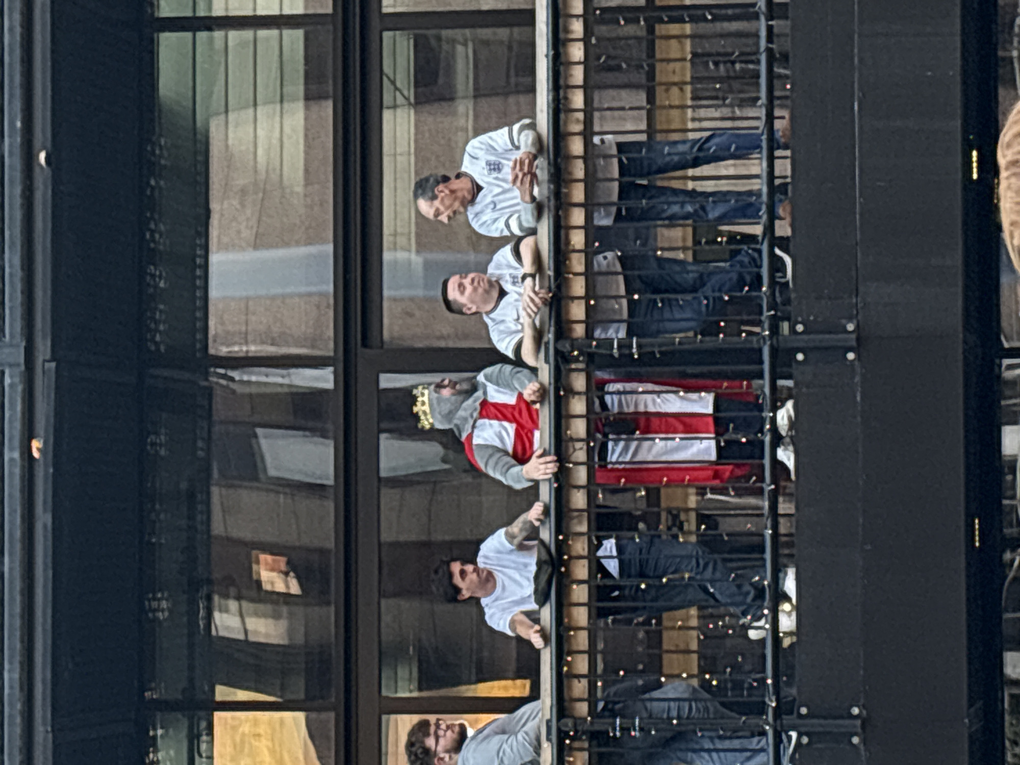 On a balcony overlooking a cityscape, five men are visible. The balcony's railing is black, and strings of tiny lights are interwoven through it. Behind the balcony is a modern building with large, reflective windows.
The men are casually dressed. One is wearing a white shirt with a red cross, a silver crown on his head, and another is in a plain white t-shirt. They appear to be looking down and interacting with something or someone out of frame. The building’s architecture and the attire suggest the scene is in London, United Kingdom, potentially during a sporting event.