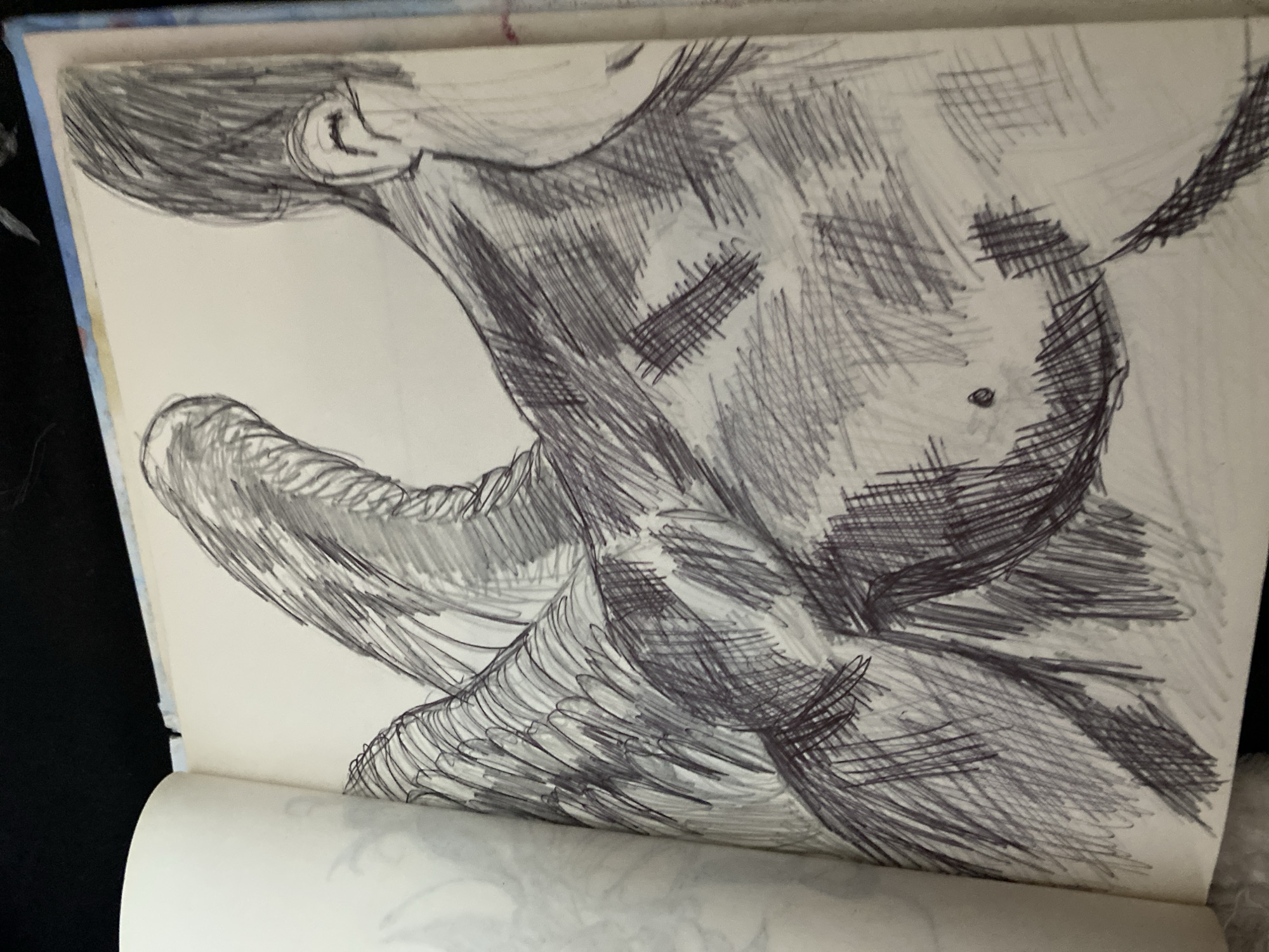 This is a close-up, angled photograph of a sketch in a notebook. The sketch is done in black ink with visible cross-hatching and directional lines used to create shading and form. The primary subject appears to be a human torso and arm, rendered in a stylized, somewhat abstract manner. The body is shown from the side, with the head and neck tilted back, revealing the ear and the curve of the skull. A substantial portion of the upper body is depicted, with musculature suggested through the heavy inking. The arm is bent, with the forearm extending forward and downward, also heavily shaded.
The sketch is on a page of what appears to be a sketchbook, with the notebook partially open, showing the binding and the curve of the pages. The background behind the sketchbook is a dark, solid color, possibly fabric, and the edge of another notebook or surface is visible at the top with a light blue and white patterned edge. There is no discernible text in the image. The lighting suggests an indoor setting, and the focus is entirely on the drawing itself, highlighting the texture and depth created by the artist's linework. The style of the sketch suggests it could be a study of anatomy or a figure drawing.