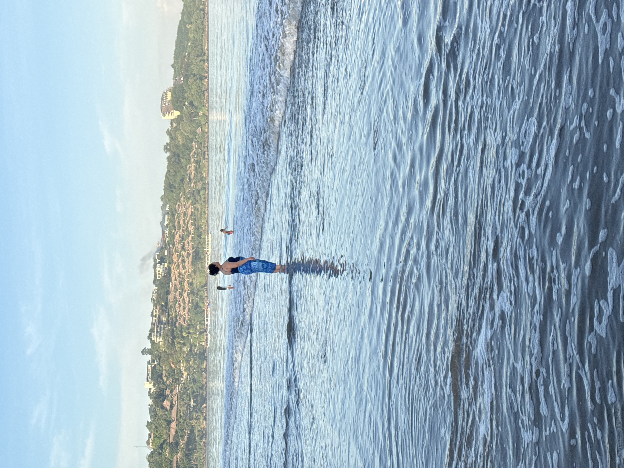 A medium, vertical shot depicts a beach scene. The view is slightly rotated counterclockwise. A woman stands in the shallow water, facing away from the camera, her body angled slightly towards the left. She has dark hair and wears a dark top and patterned blue shorts. The water is clear and ripples around her legs, creating a reflection. Other individuals are visible in the distance, further along the shoreline.
The water transitions from the light blue near the shore to a darker hue further out. Waves are visible in the middle ground. The sky is clear blue with light clouds near the horizon.
In the background, a coastline is lined with trees and buildings. It features a mix of residential and commercial structures, including what appear to be hotels or resorts. Based on the buildings, location, and appearance of the beach, the scene is likely in Jimbaran, Indonesia.