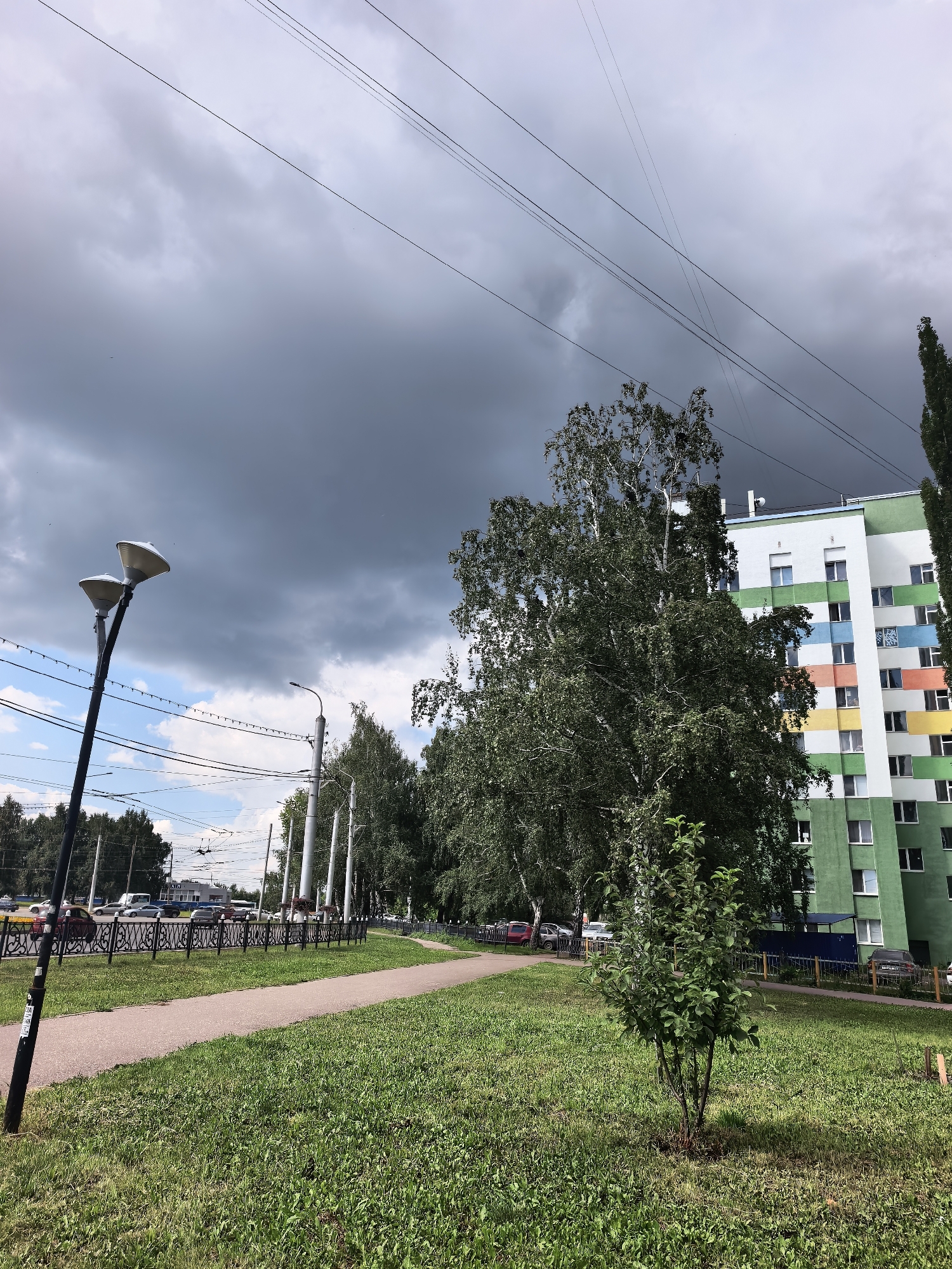 The image captures a street scene in Sterlitamak, Russia, on a partly cloudy day. A paved walkway runs through a grassy area, bordered by a decorative metal fence. Beyond the fence, a street with moving vehicles and trolleybus wires is visible. A tall apartment building, painted with green and multi-colored horizontal stripes, stands on the right side of the frame. Several trees, including a large birch tree, are situated in the foreground and mid-ground. Two streetlights are present on the left side of the walkway. The sky is a mix of dark storm clouds and lighter blue patches.